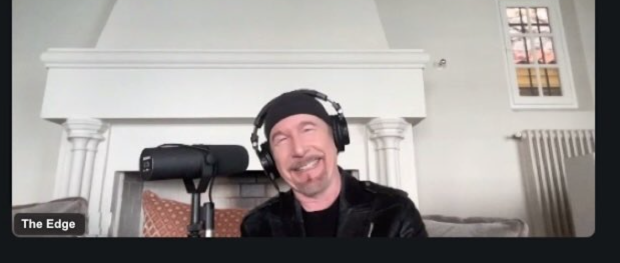 A screenshot of a Zoom call showing U2’s The Edge smiling in a room with headphones on and a Shure SM7B microphone in front of him.