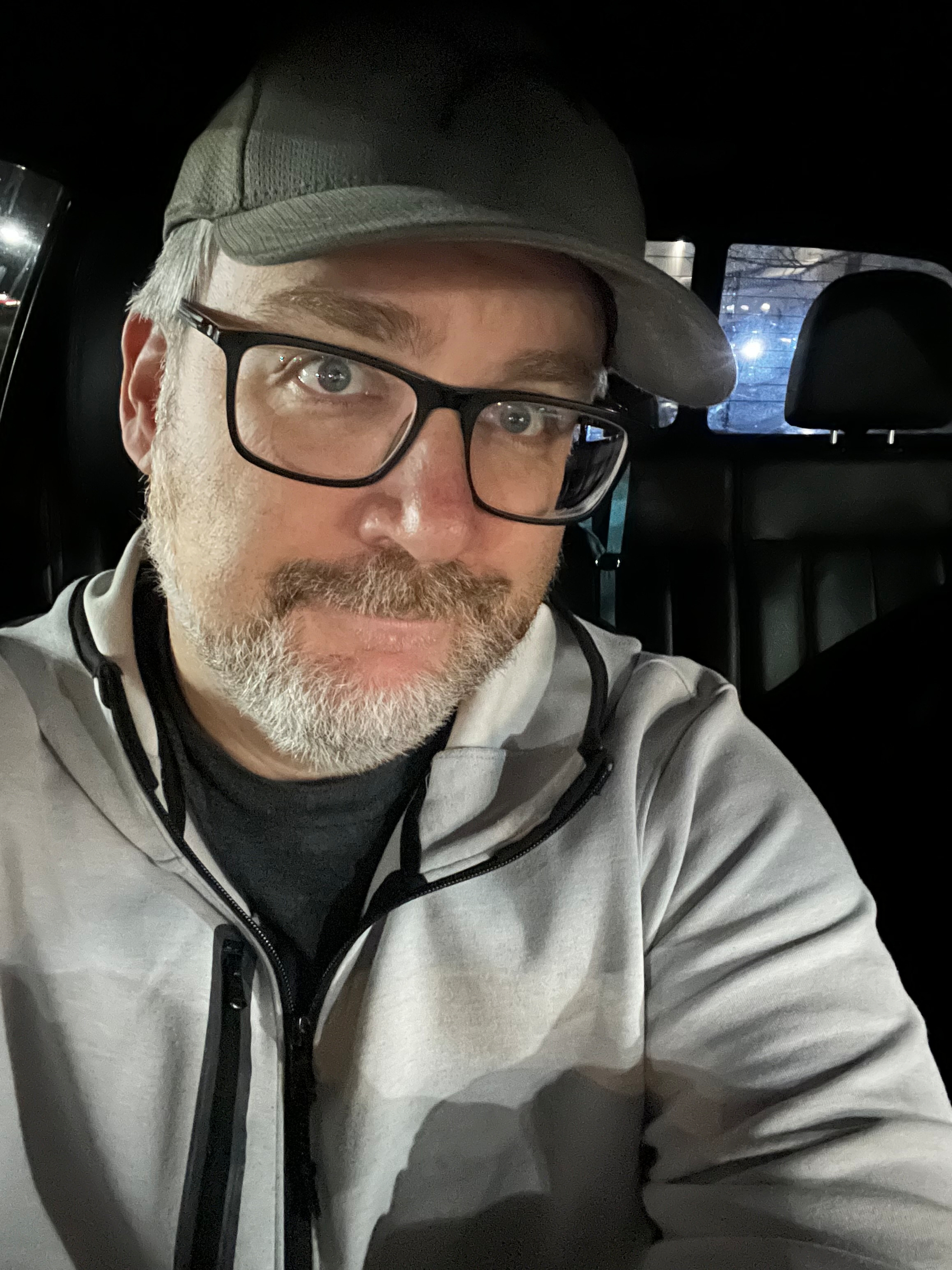 A person wearing glasses and a cap is seated inside a vehicle, with a slight smile and ambient lighting.