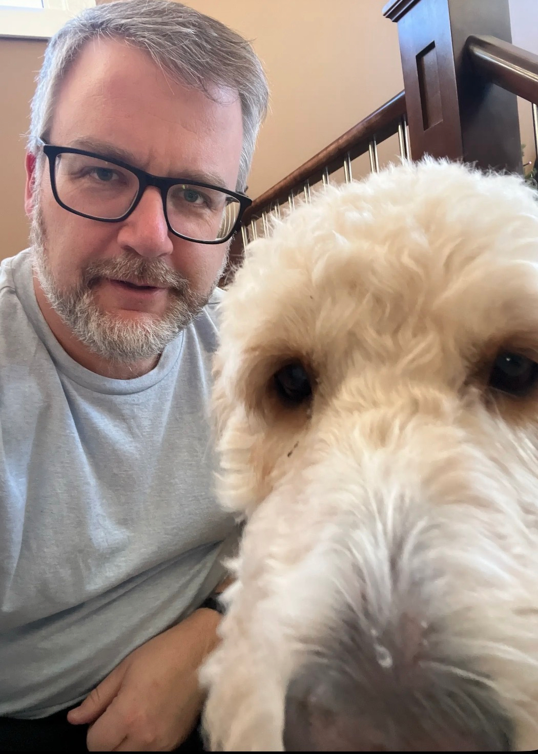 A man with glasses and a beard poses next to a fluffy dog indoors.