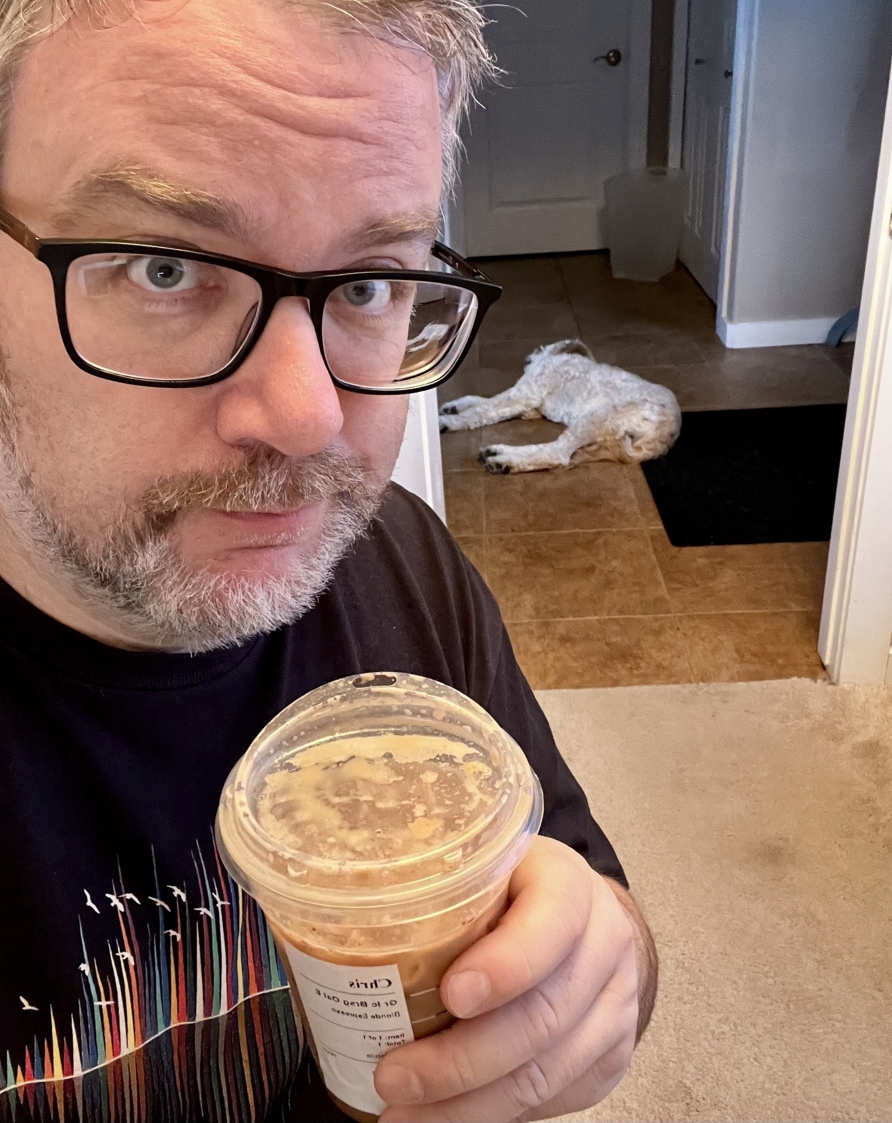 A person with glasses holds a iced beverage, while a dog lies on the floor in the background.