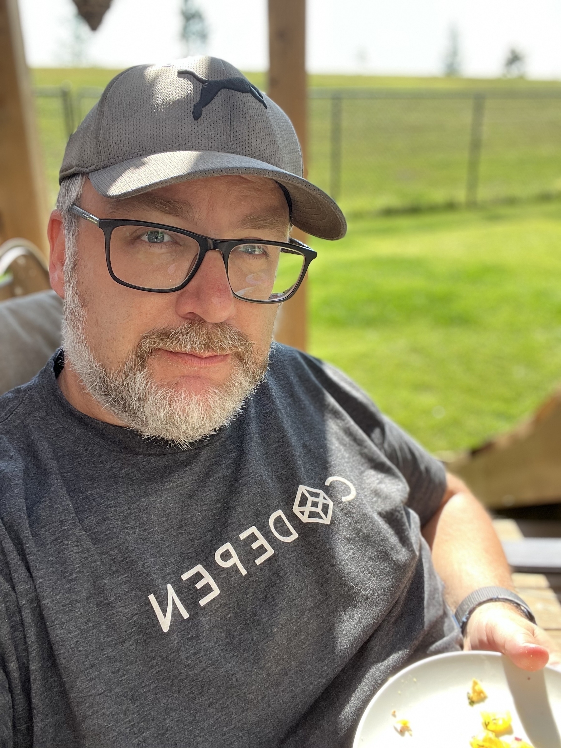 A man wearing glasses and a cap sits outdoors, holding a plate with a small amount of food.