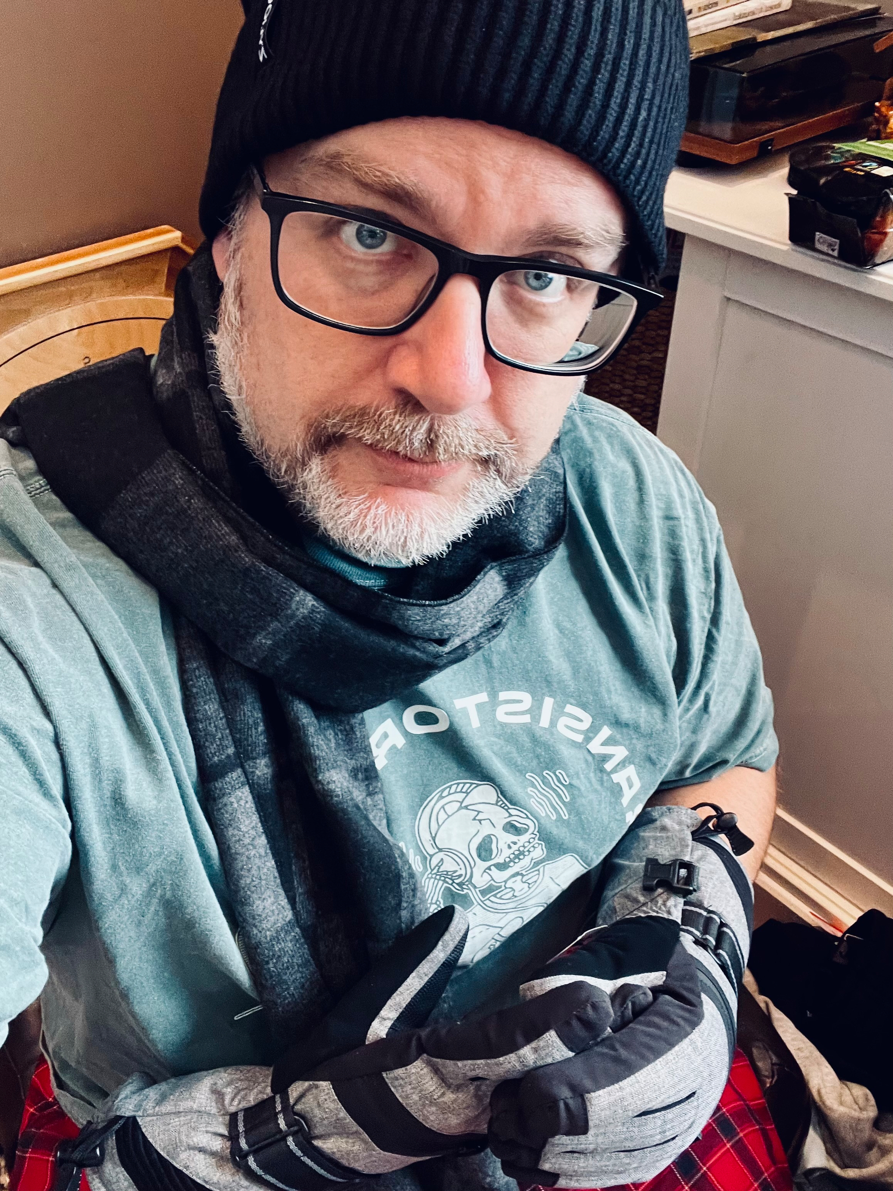 A person wearing glasses, a beanie hat, a scarf, and gloves poses while sitting indoors.