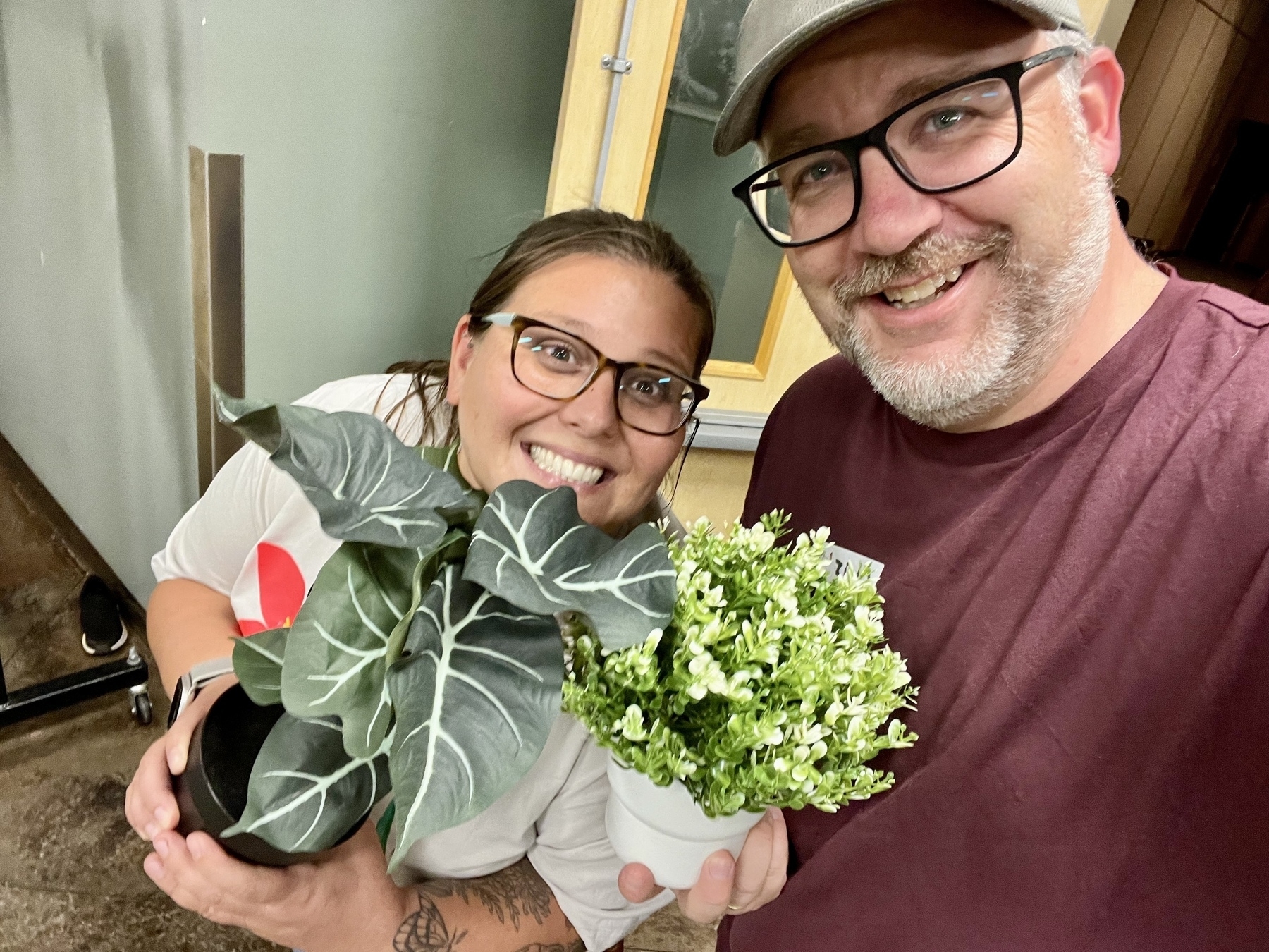 Two people smiling while holding potted plants indoors.