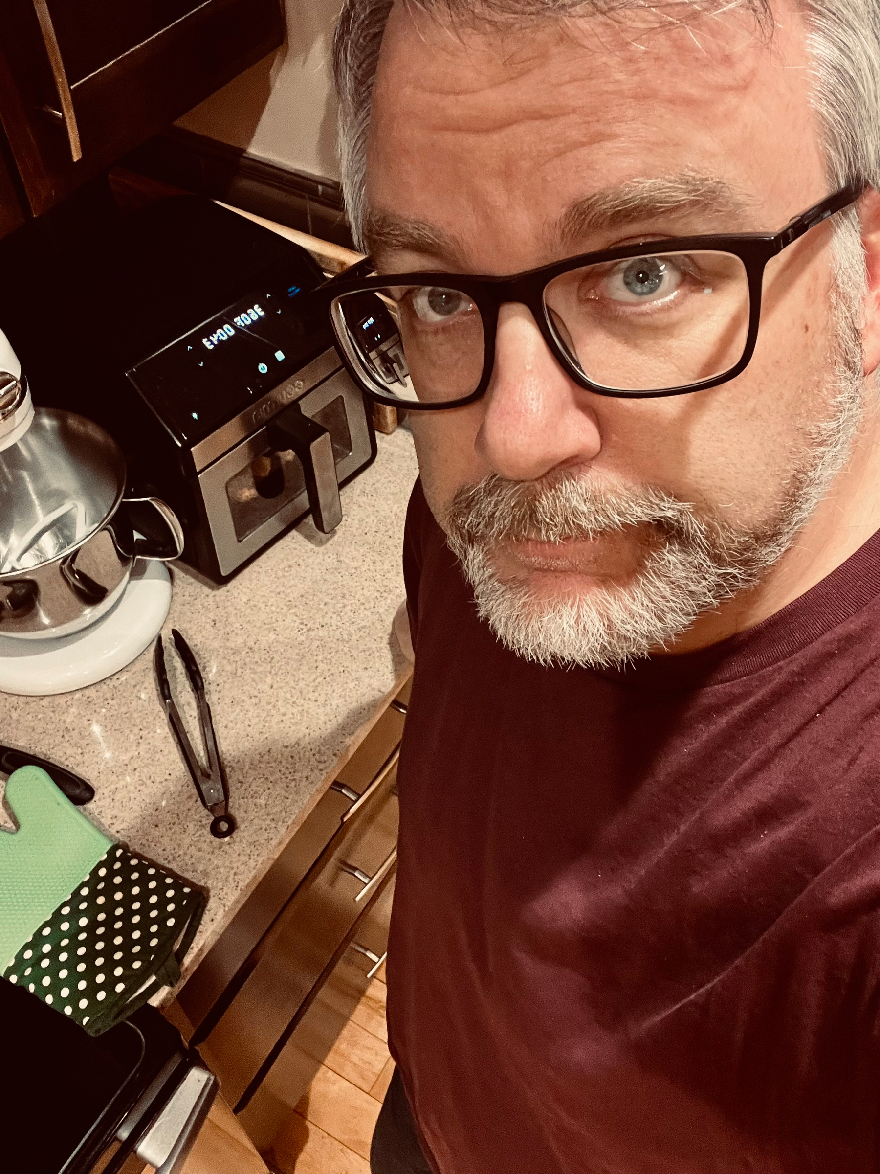 A person with glasses and a beard is standing in a kitchen near a countertop with a toaster oven, mixer, and kitchen towel.