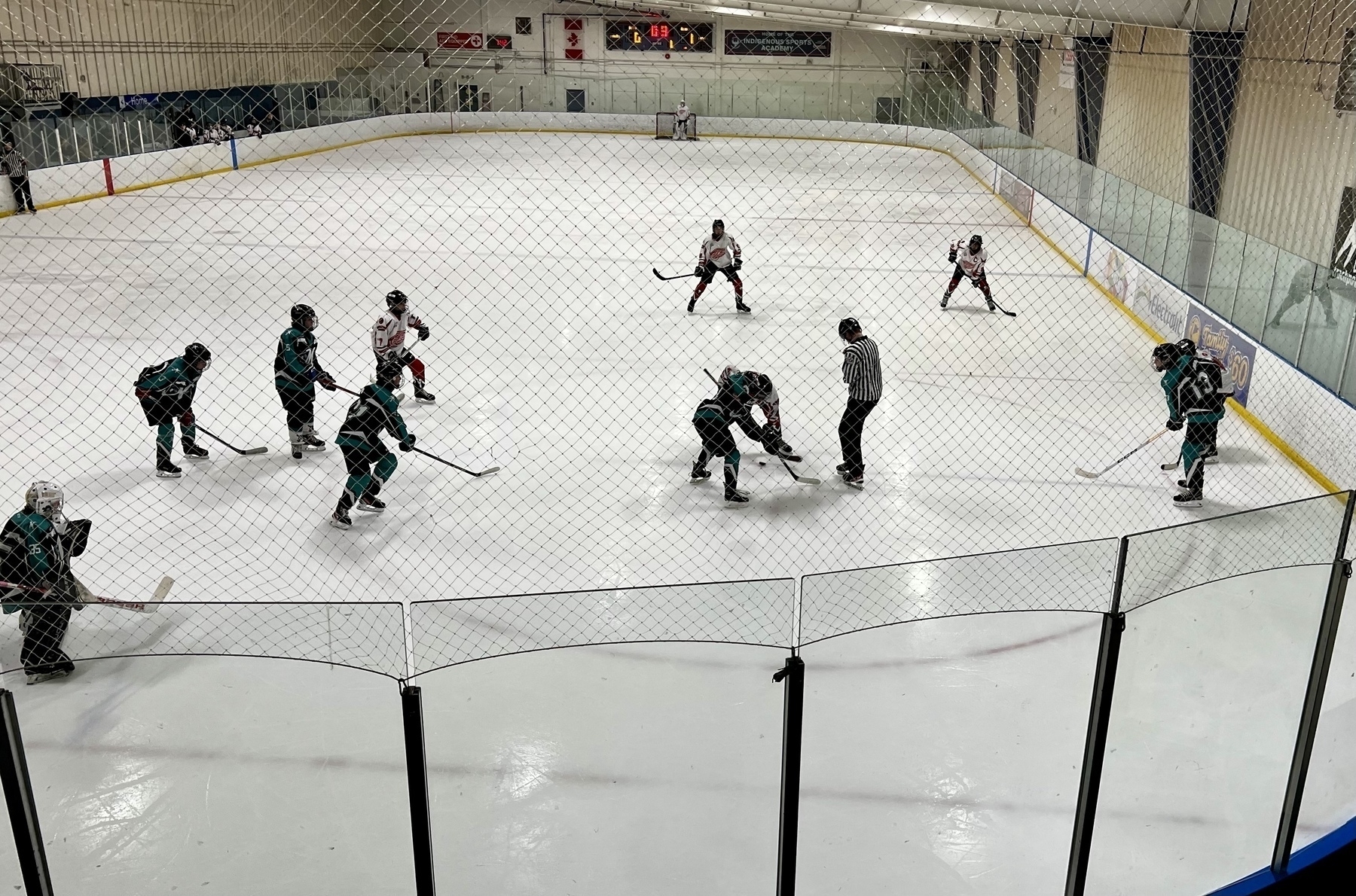 View of a minor hockey game rink with the players just starting play at a face-off near the goalie. 