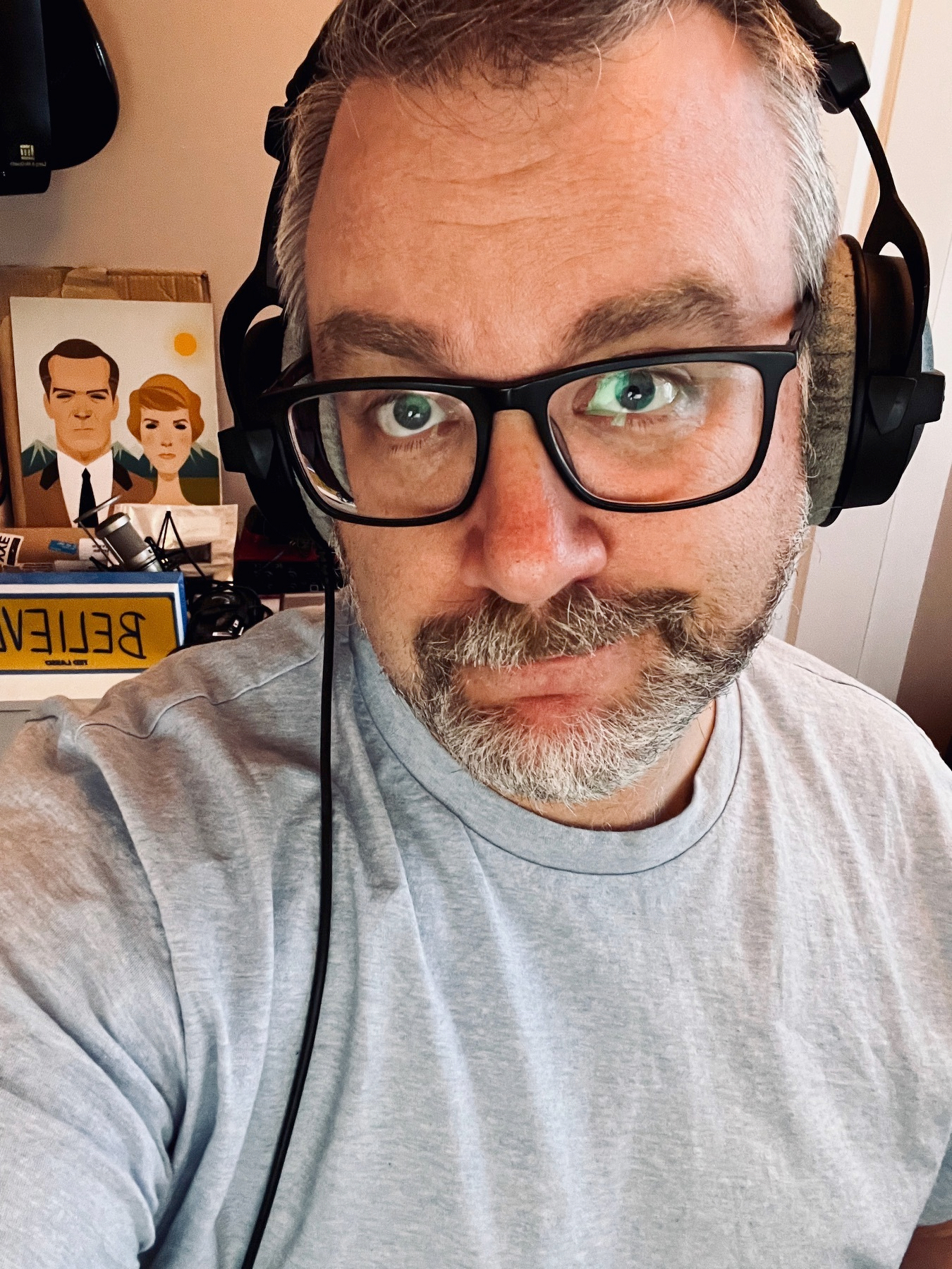 A bearded man wearing headphones and glasses takes a selfie in an indoor setting.