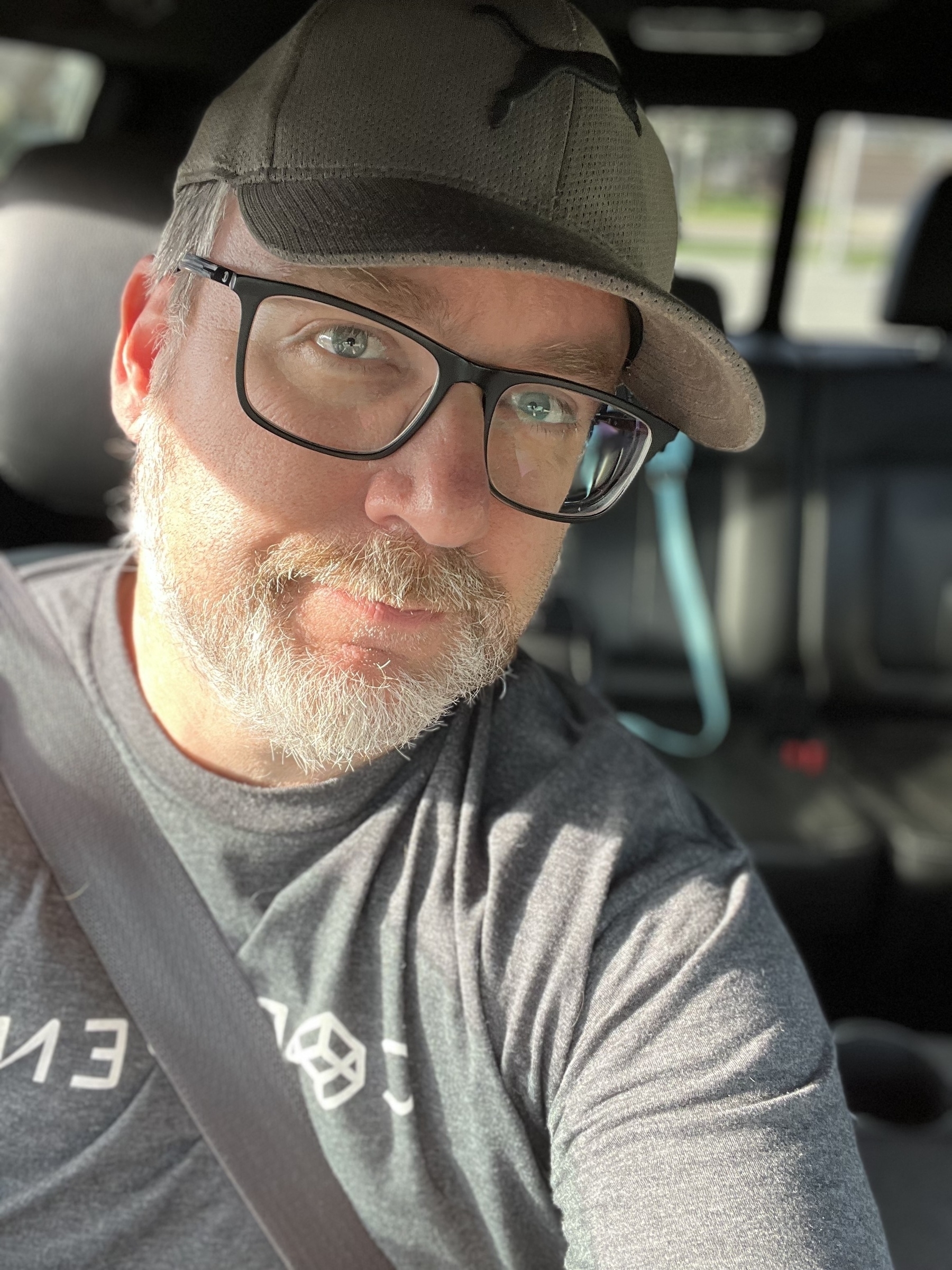 A person wearing glasses and a cap is seated in a car, looking towards the camera.
