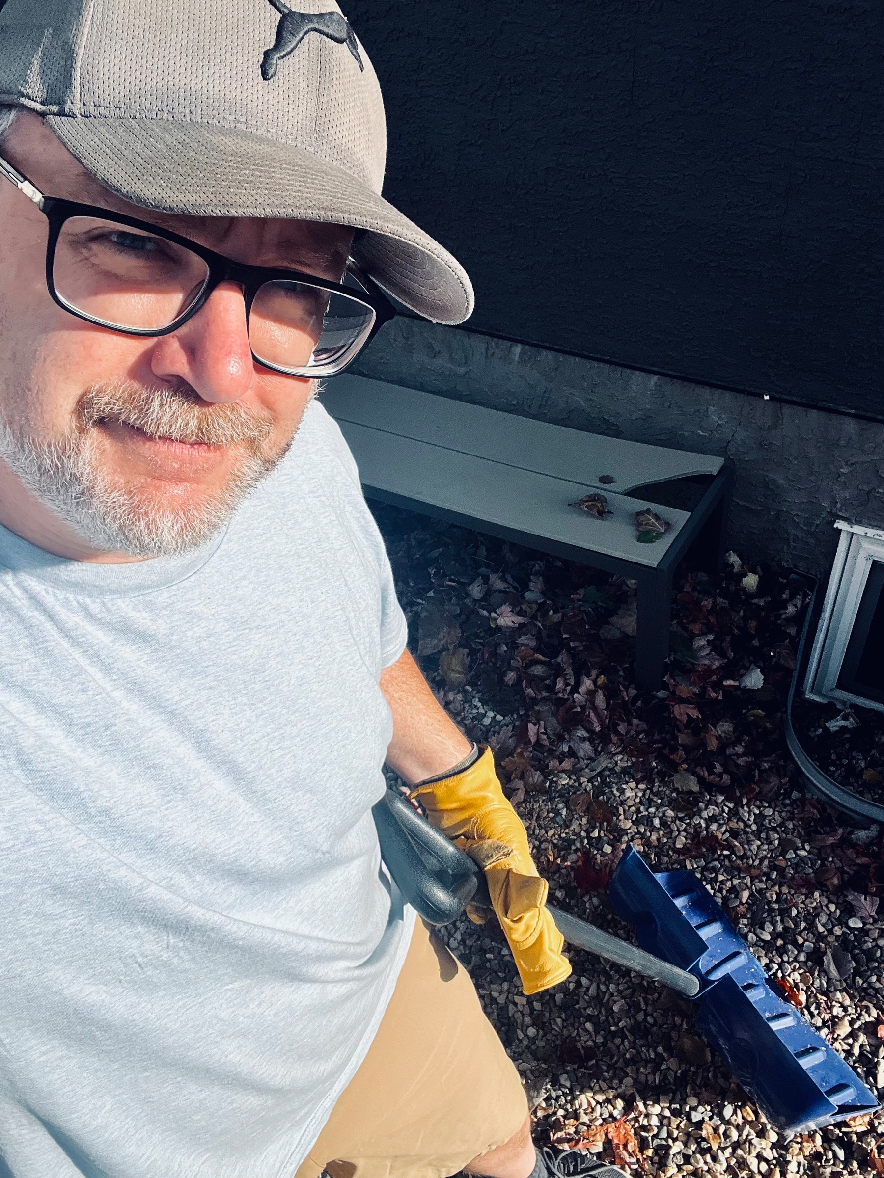 A person wearing a cap and glasses is holding a blue rake on a pebble and leaf-covered ground.