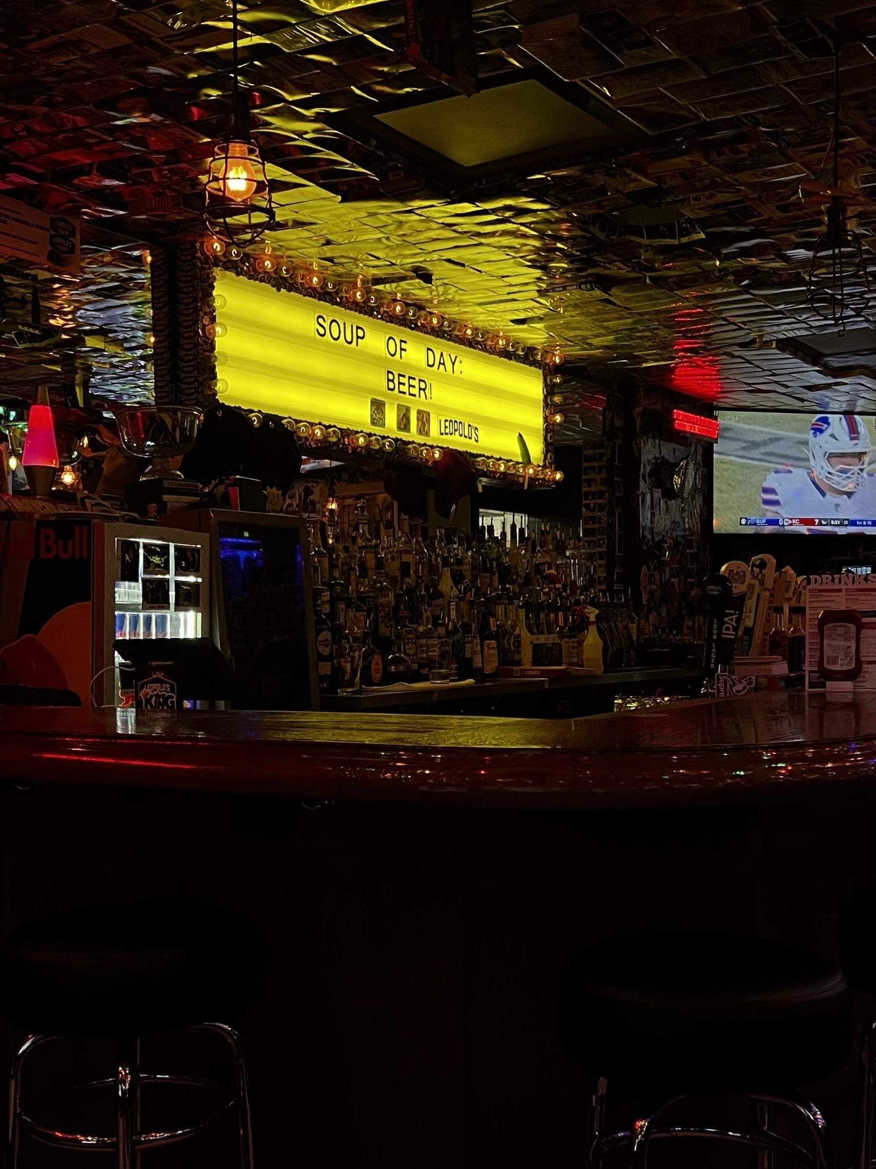 A dimly lit bar features a humorous sign reading "SOUP OF DAY: BEER!" and a television showing a sports game.