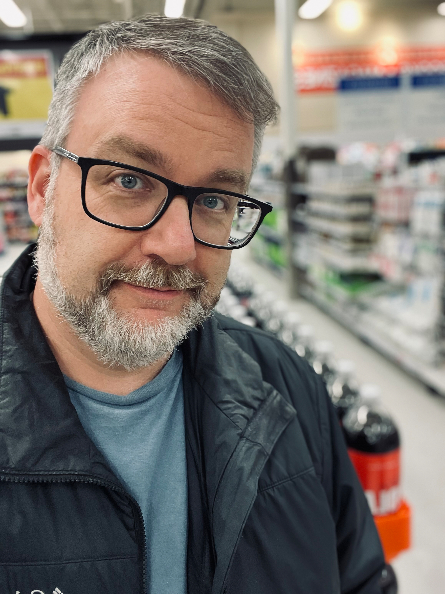 A person with glasses and a beard is standing in a store aisle filled with beverages.