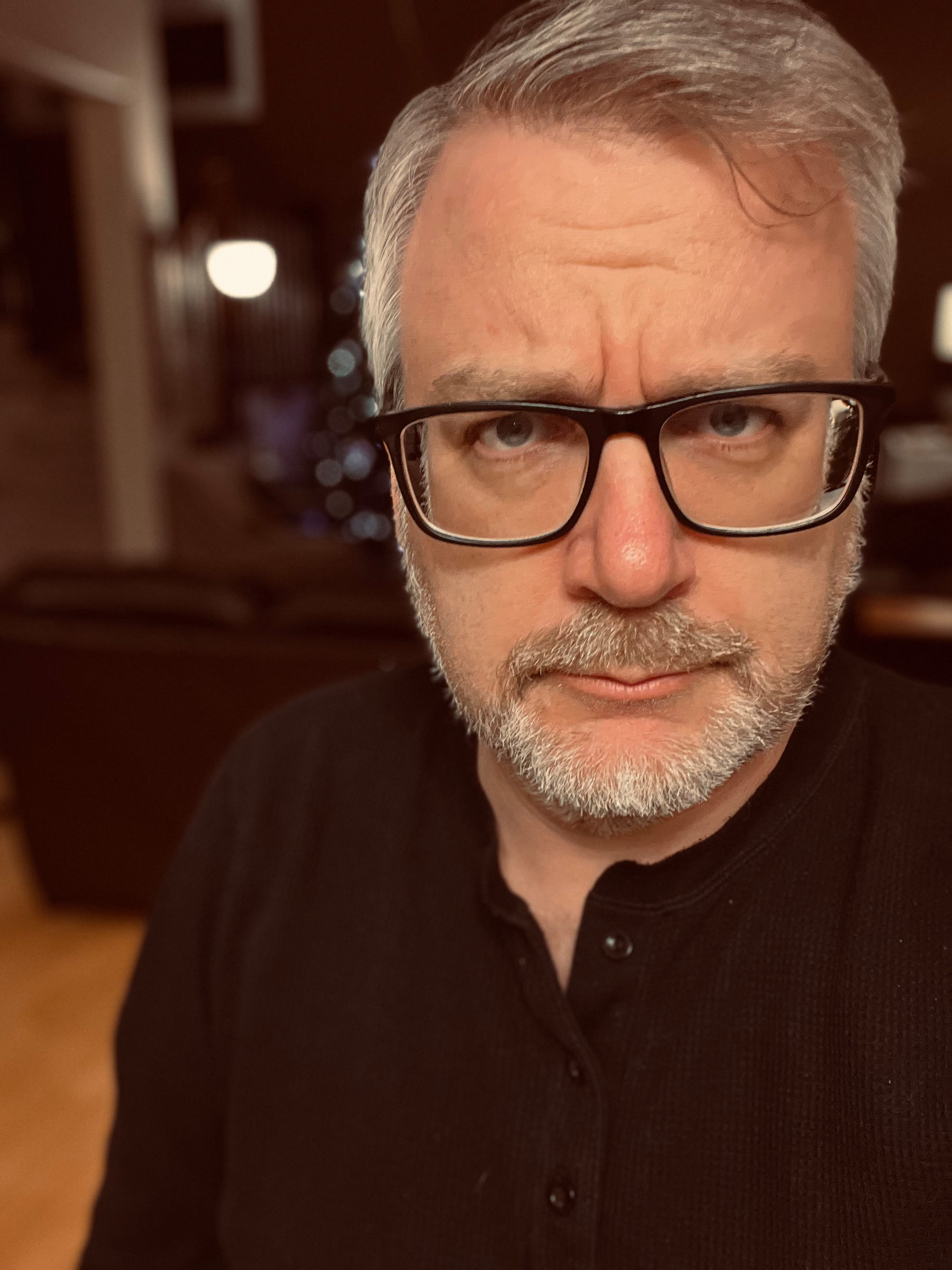 A man with glasses and gray hair is looking directly at the camera with a slightly raised eyebrow.
