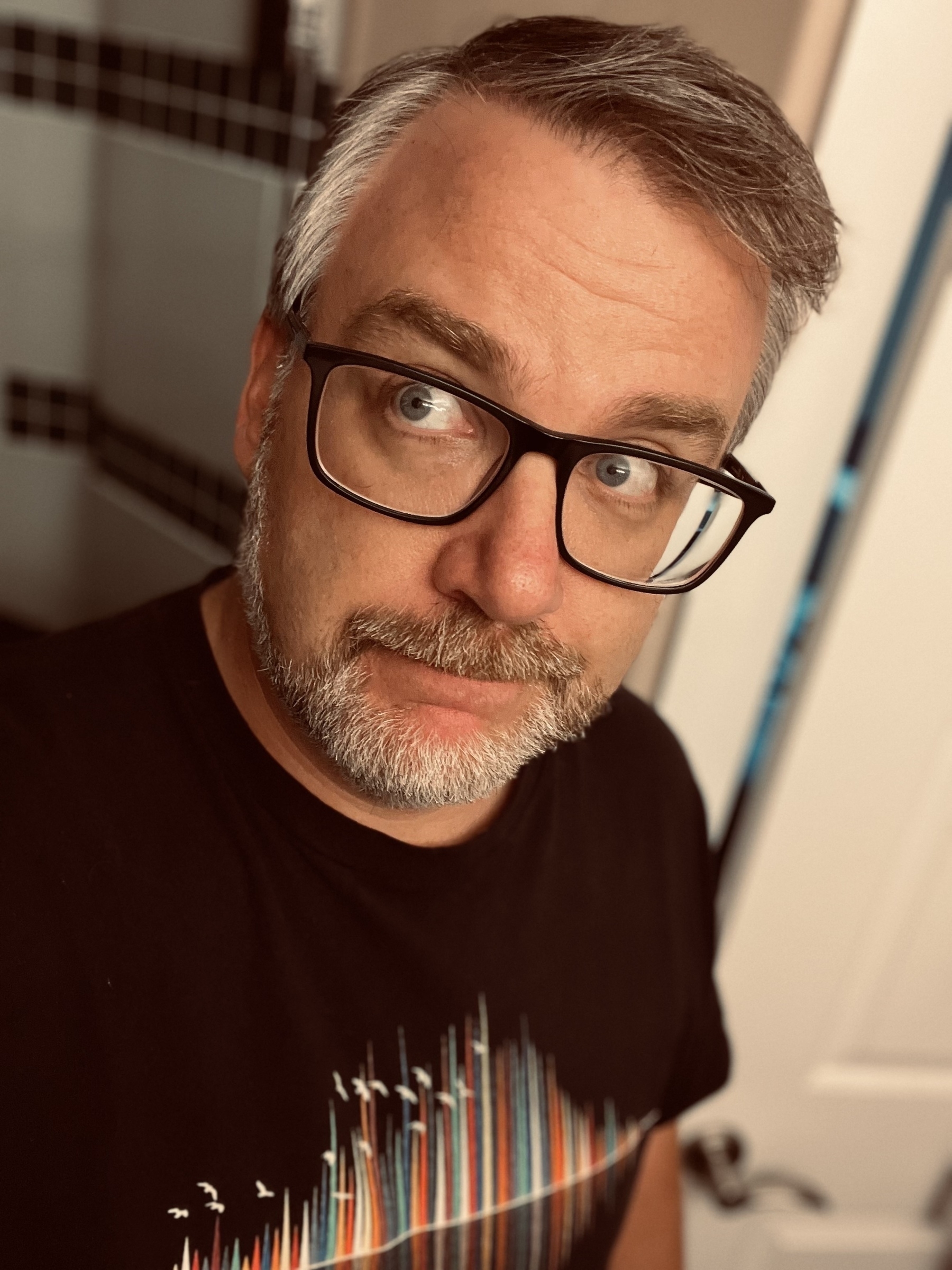 A person with glasses and a beard is wearing a black shirt featuring a colorful design.