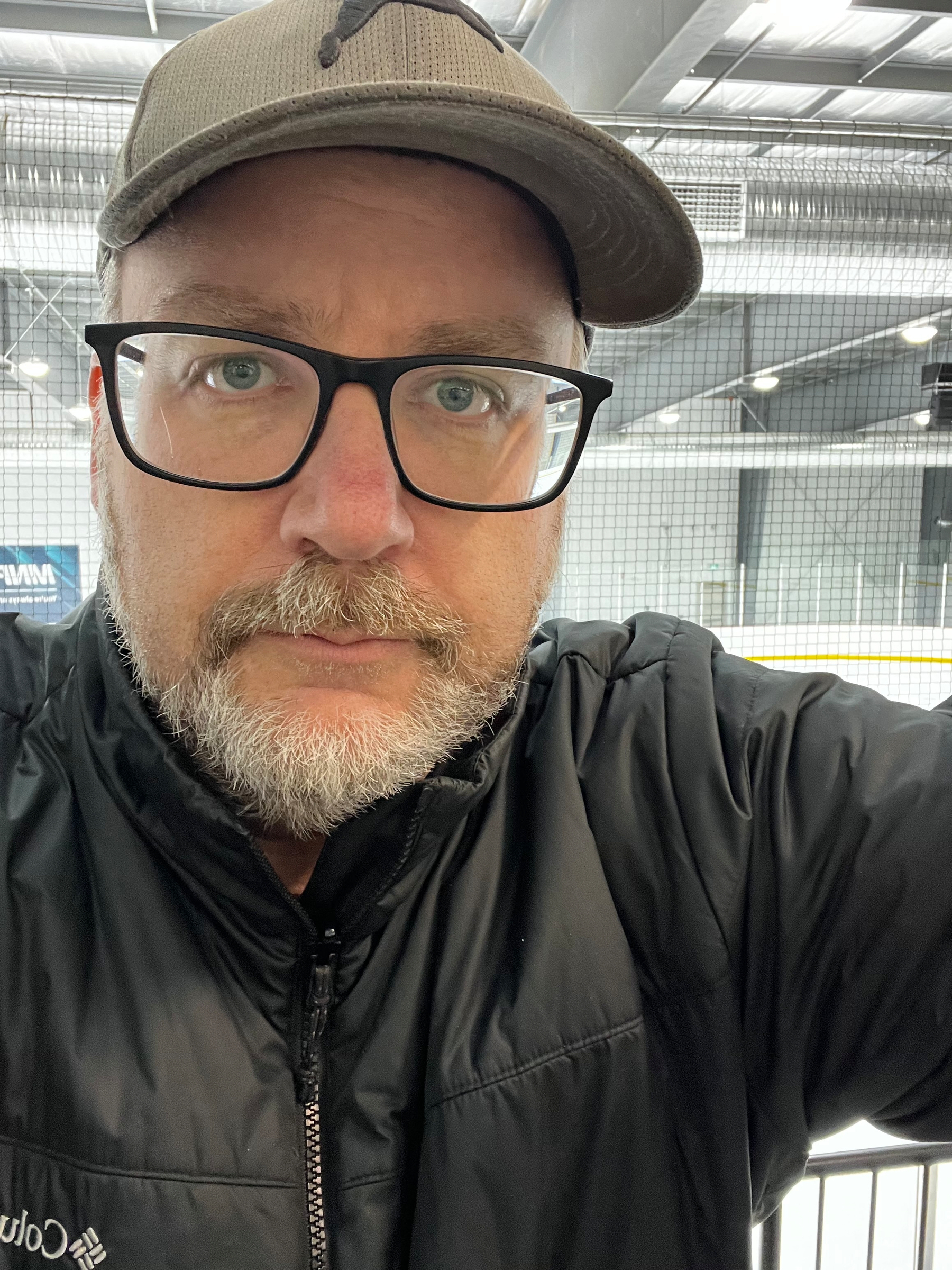 A person wearing glasses and a cap stands indoors, possibly near an ice rink.
