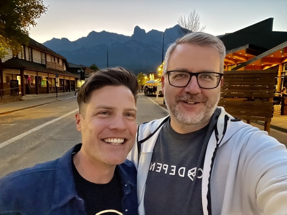 Two people are smiling for a selfie on a street with mountains in the background at dusk.