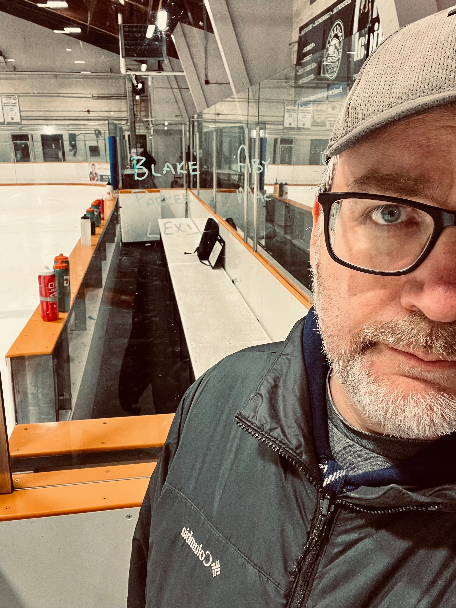 A person wearing glasses and a cap stands next to an ice hockey rink with benches and gear nearby.