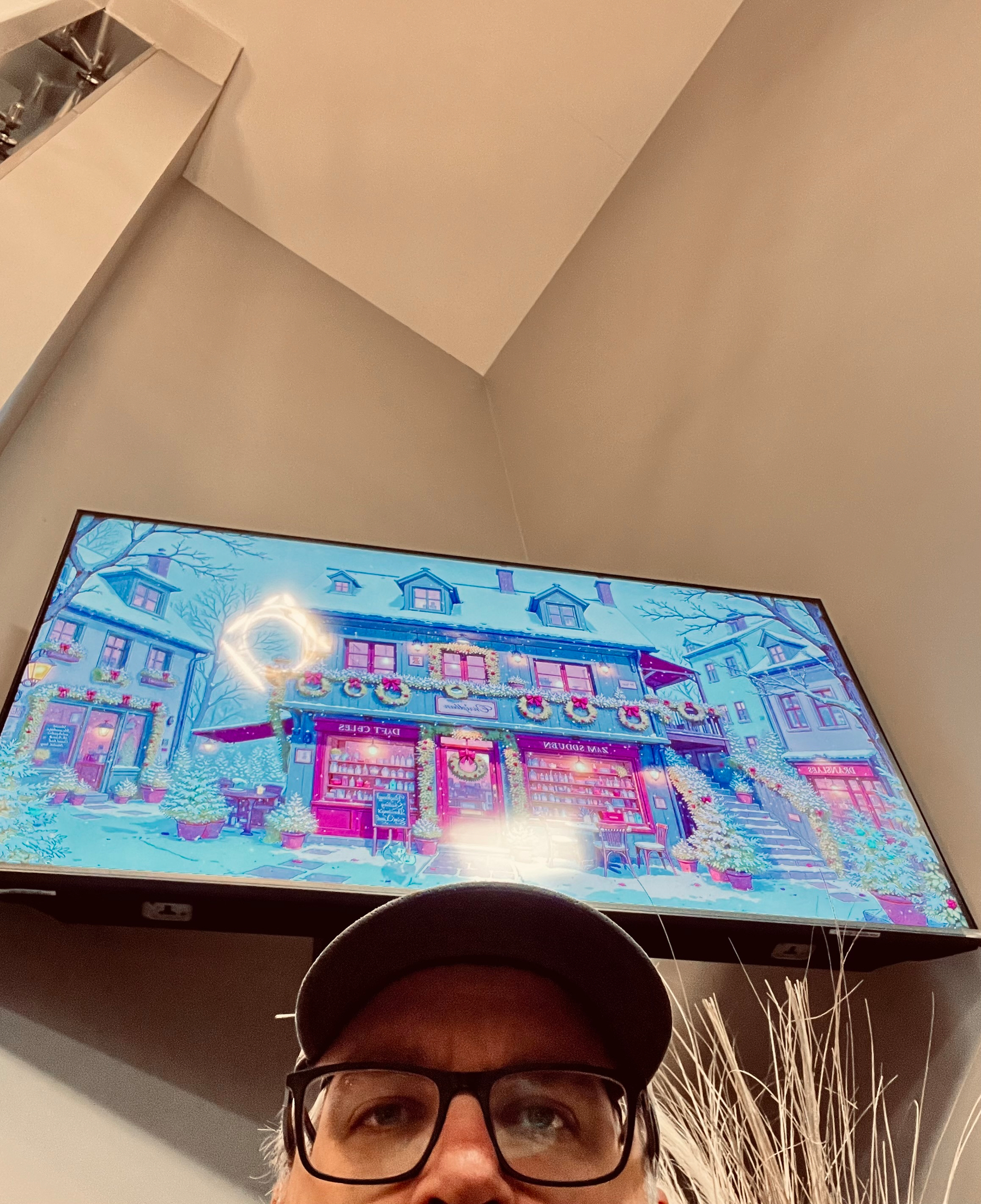 A person wearing glasses and a cap is taking a selfie in front of a screen displaying a colorful digital illustration of a quaint building with shops.