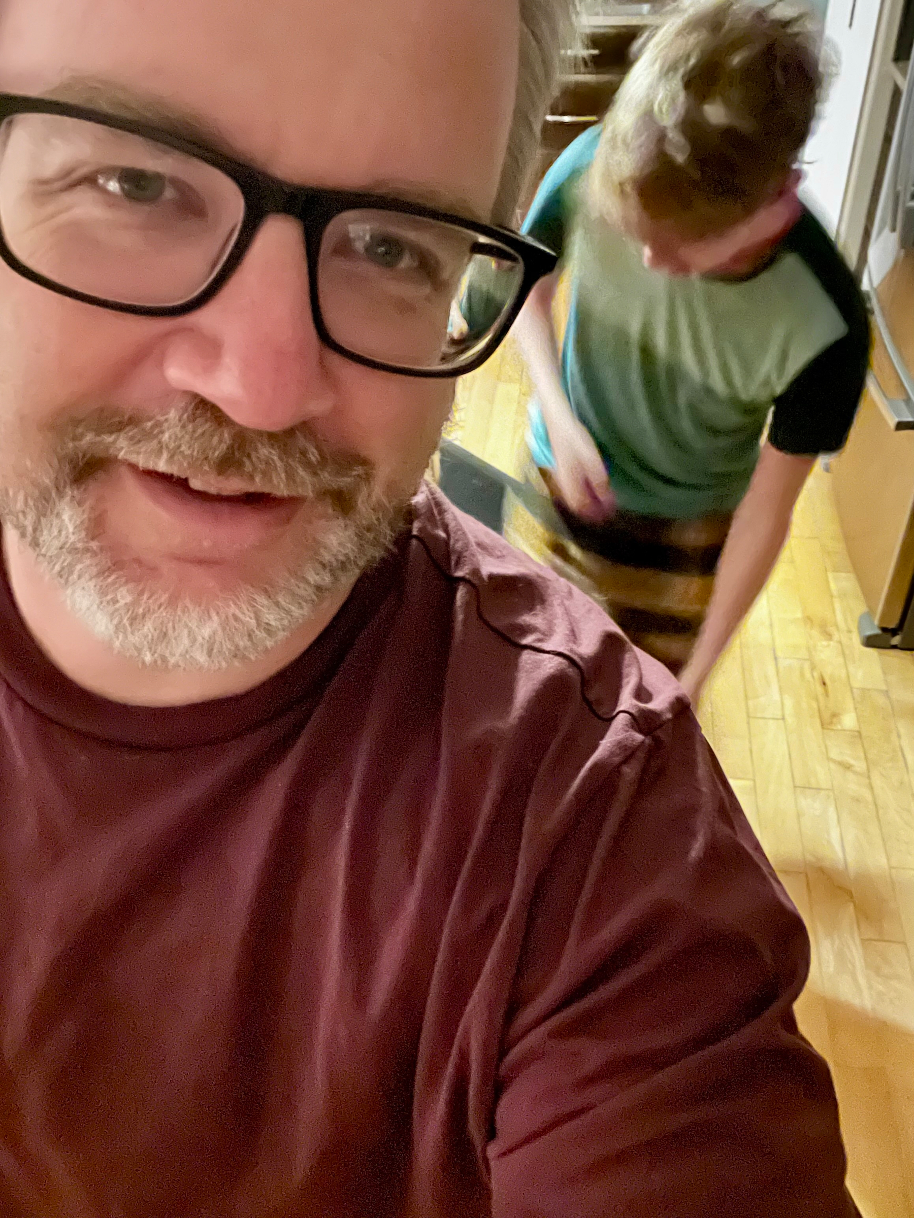 A man with glasses smiles at the camera while another person is blurred in the background.