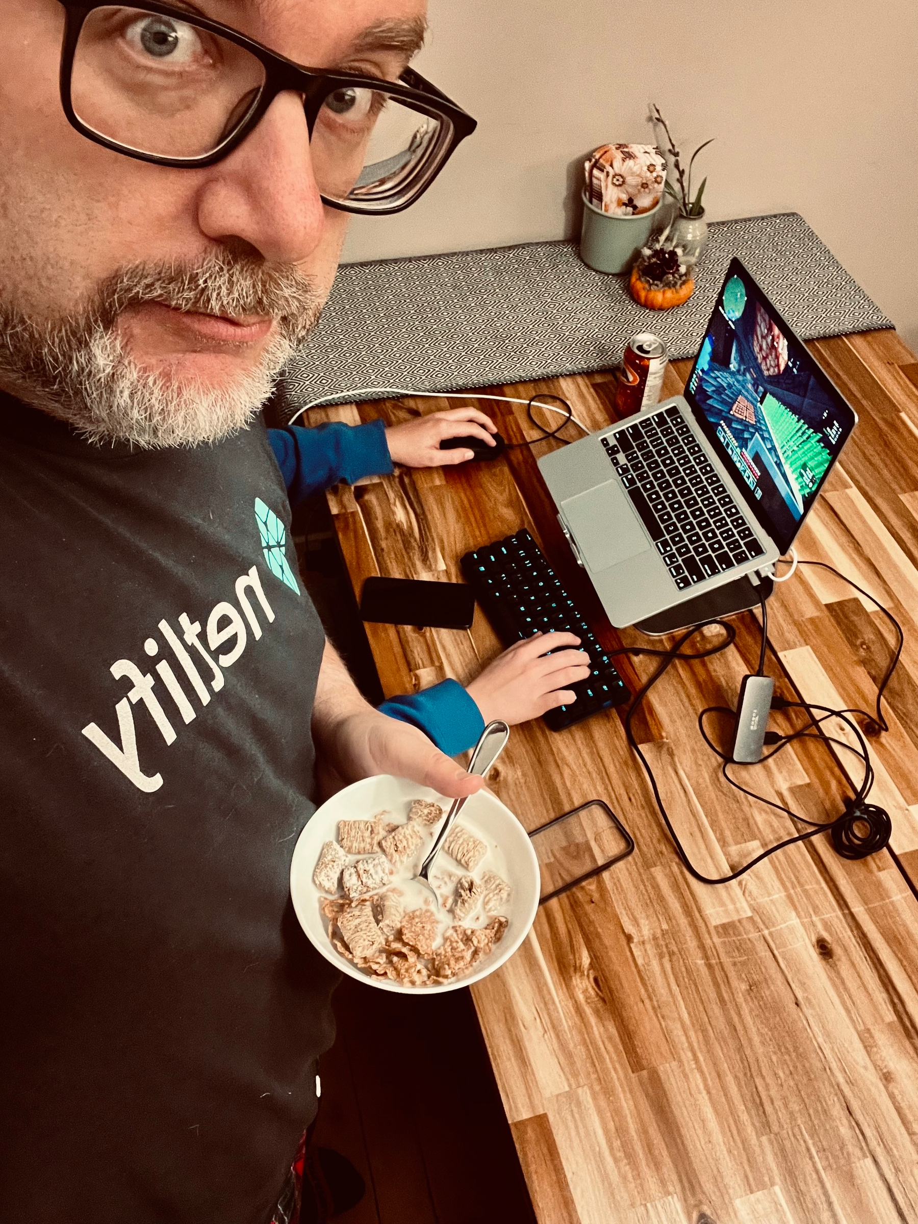 A person is holding a bowl of cereal while using a laptop on a wooden table, surrounded by various gadgets and a drink can.