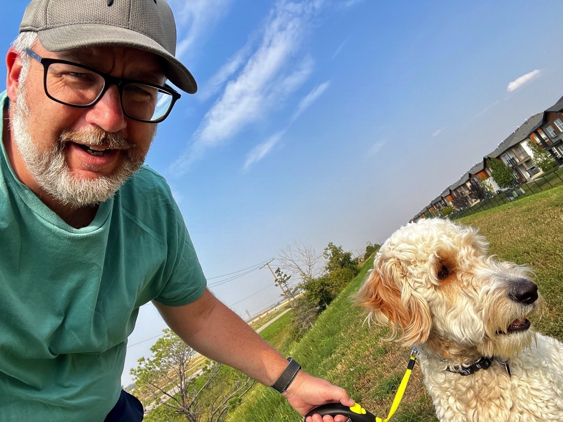 Selfie of me, a man with a hat, glasses, and a beard, bent over to include a golden doodle puppy named Neville Dogbottom in the photo. We’re outside on a walk on a sunny, fall day.
