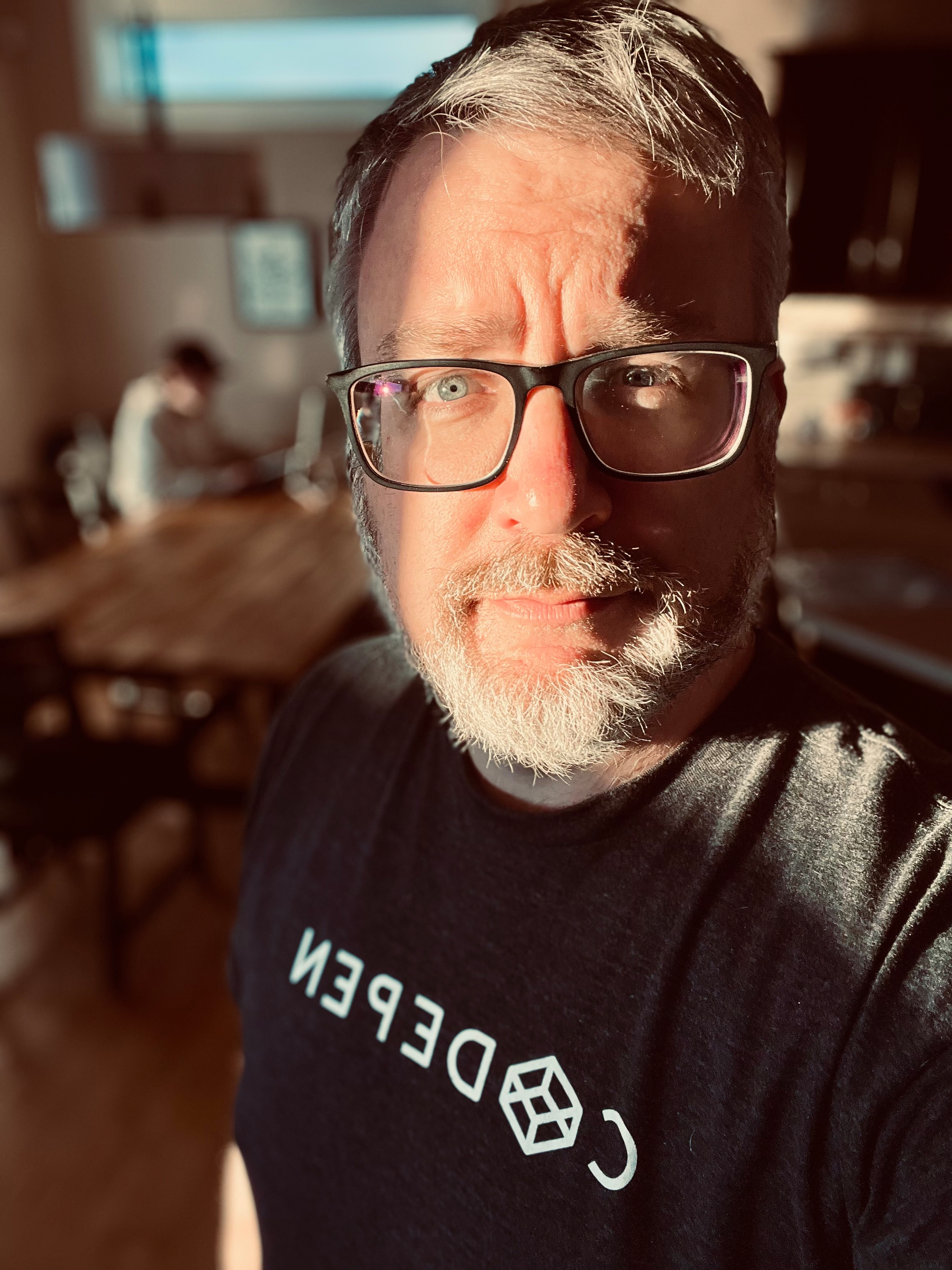 A bearded man with glasses is taking a selfie in a warmly lit room, wearing a shirt with text and a person seated in the background.