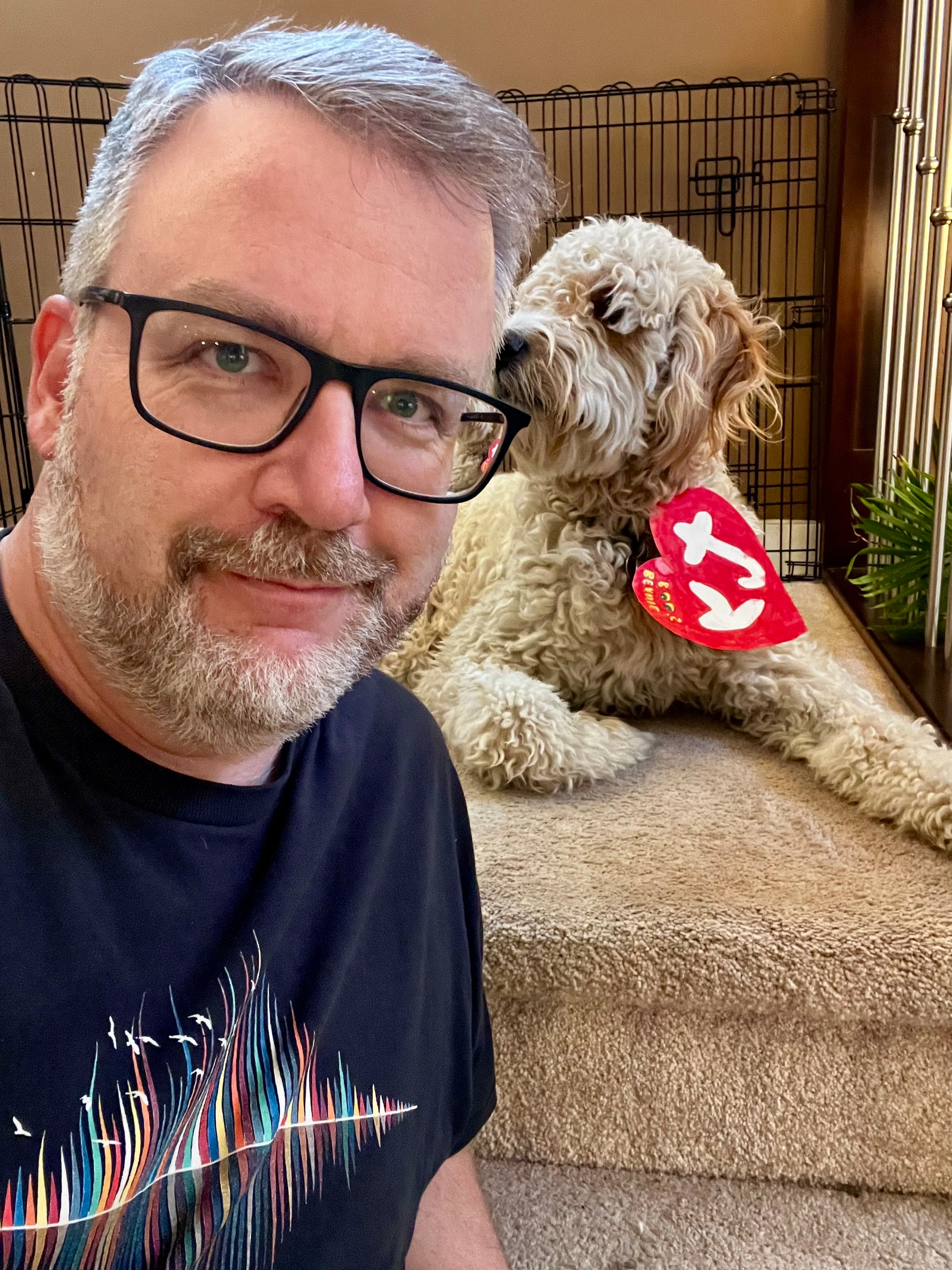 A man wearing glasses poses for a selfie with a fluffy dog that has a tag resembling a Beanie Baby's heart-shaped label.