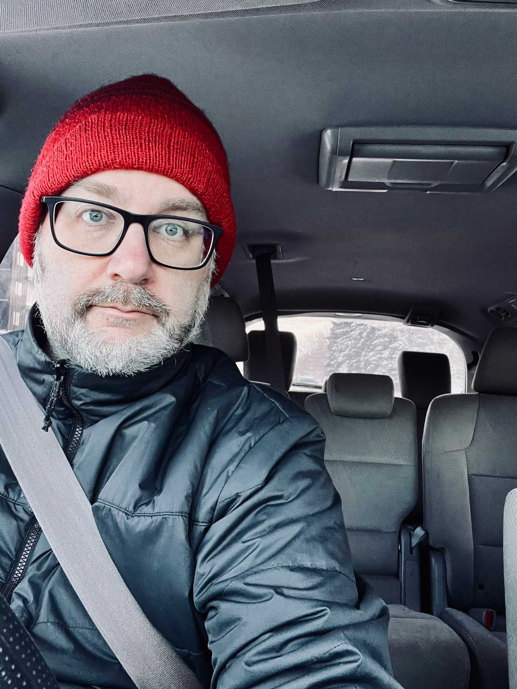 A person wearing a red beanie and glasses is sitting in the driver's seat of a car, looking at the camera.