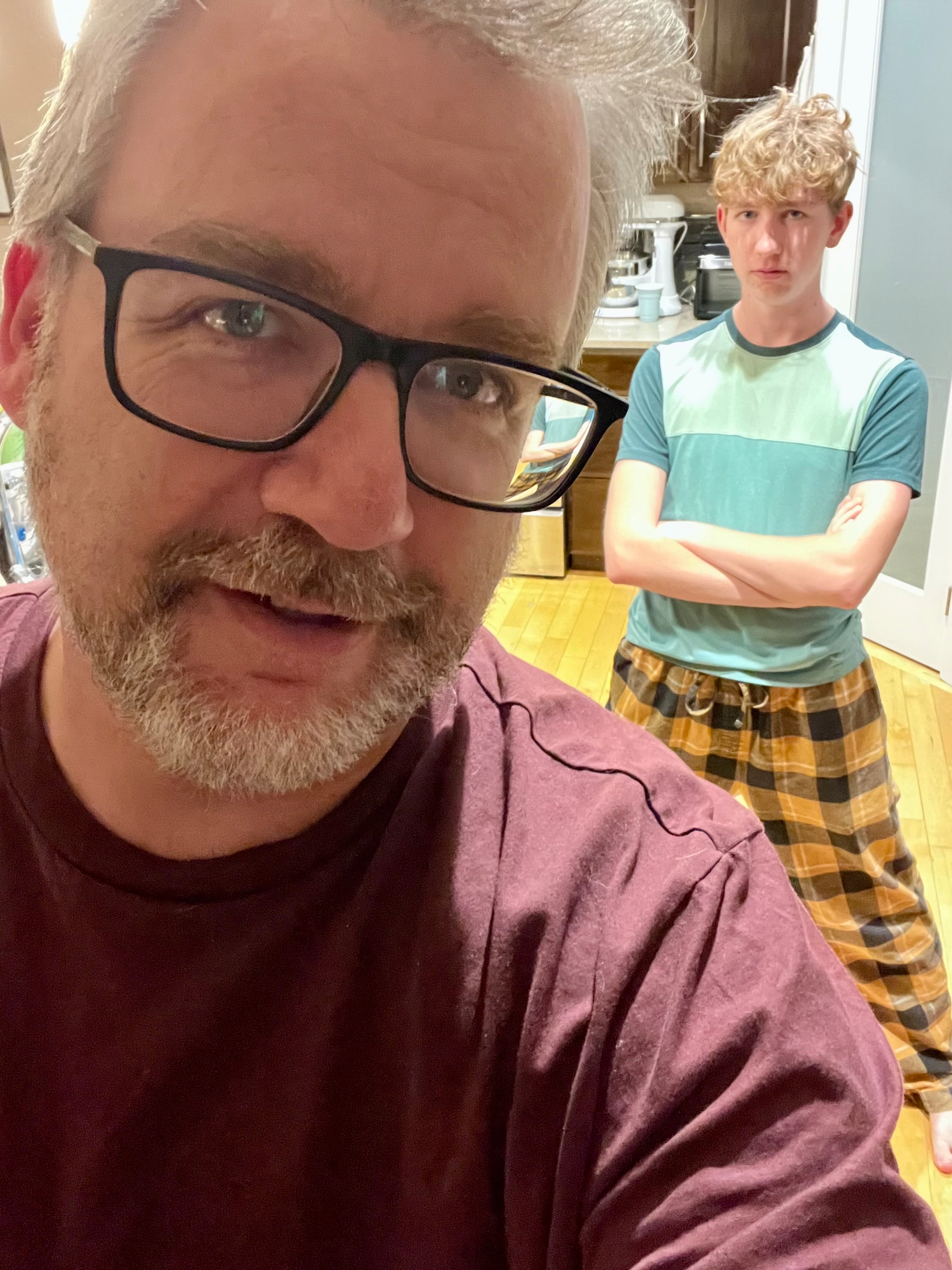 A man wearing glasses takes a selfie with a younger person in a blue and green shirt and checkered pants standing in the background with crossed arms.