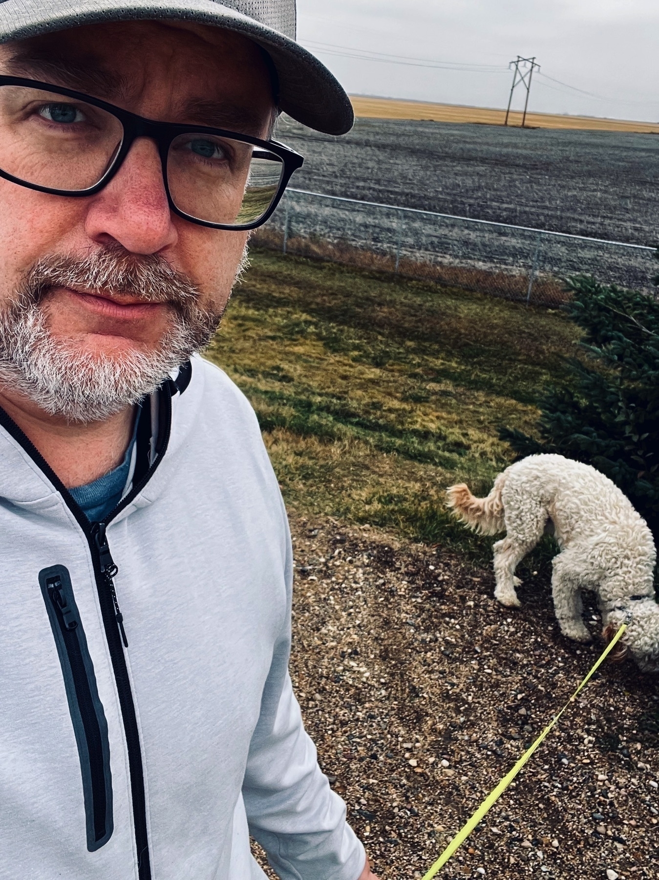 A man wearing glasses and a cap stands outdoors with a leashed dog sniffing the ground.