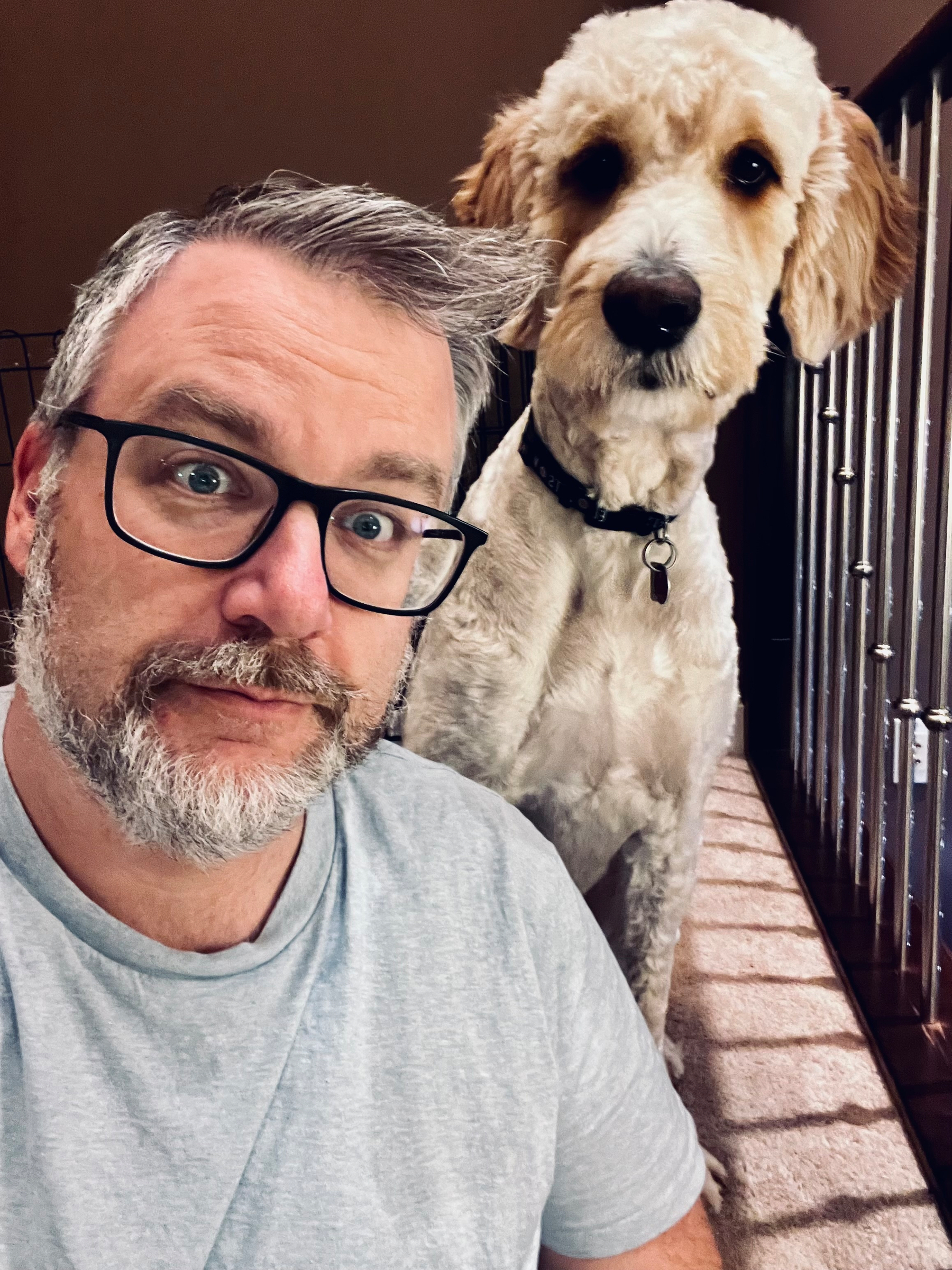 A man with glasses and a gray beard poses with a fluffy dog beside him indoors.