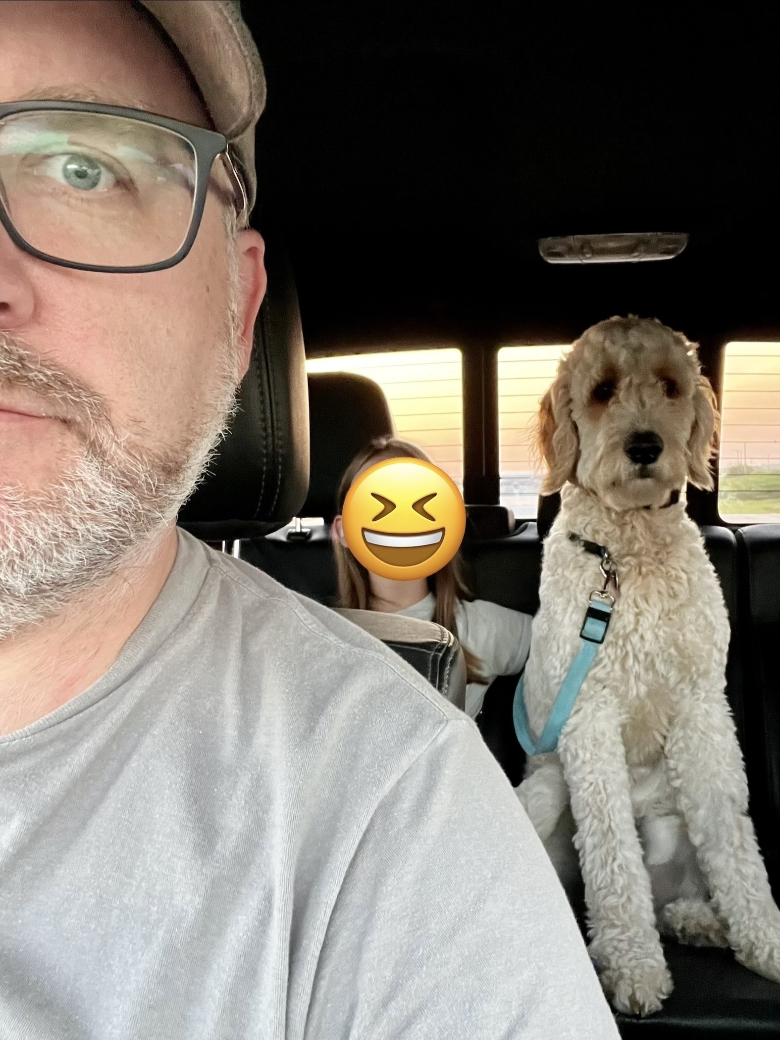 A man wearing glasses and a hat is in the front seat of a car, with a dog sitting in the back next to a child whose face is covered by an emoji.