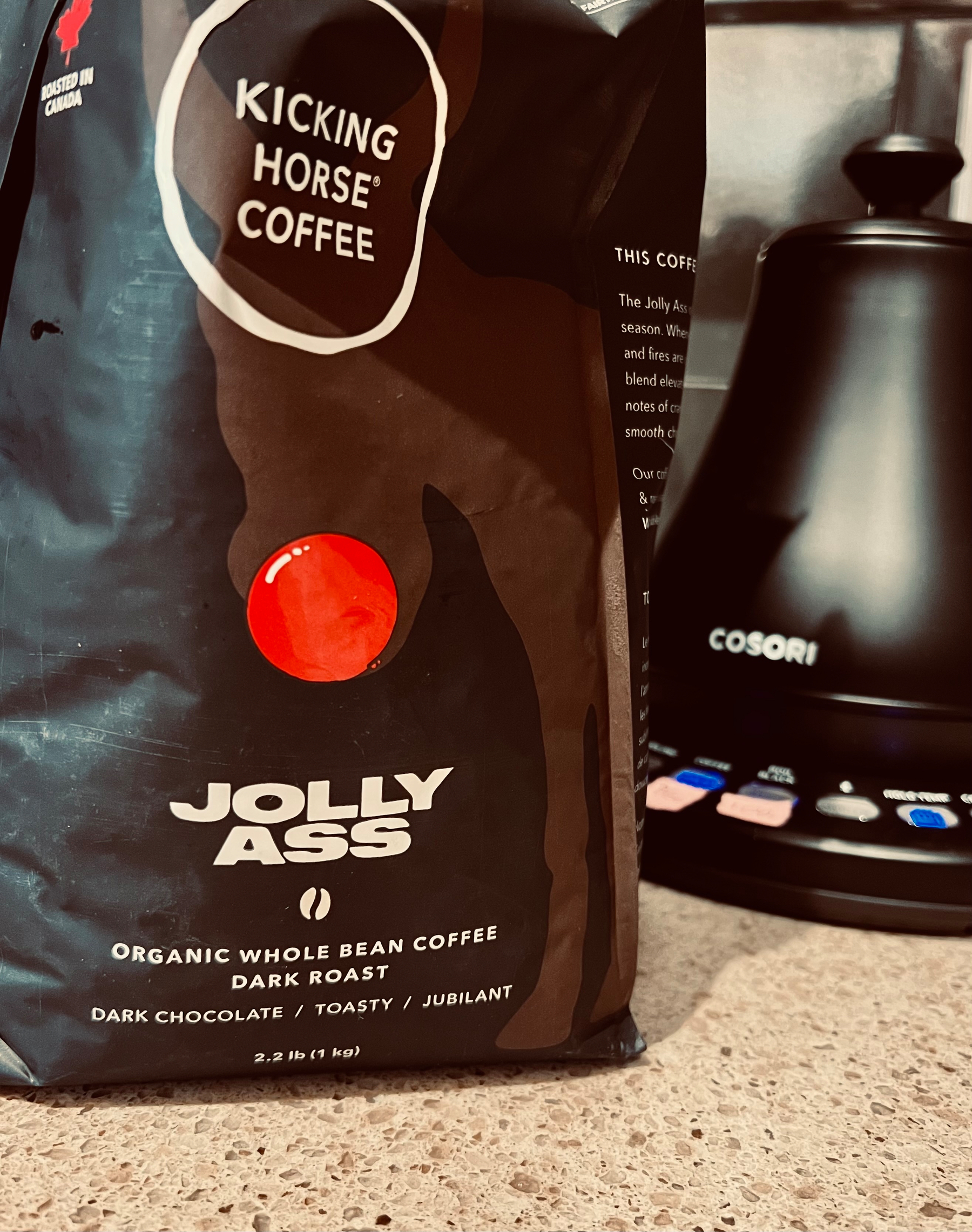 A bag of Kicking Horse Coffee labeled Jolly Ass, featuring dark roast organic whole bean coffee with tasting notes of dark chocolate, toasty, and jubilant, is placed on a countertop next to a Cosori appliance.