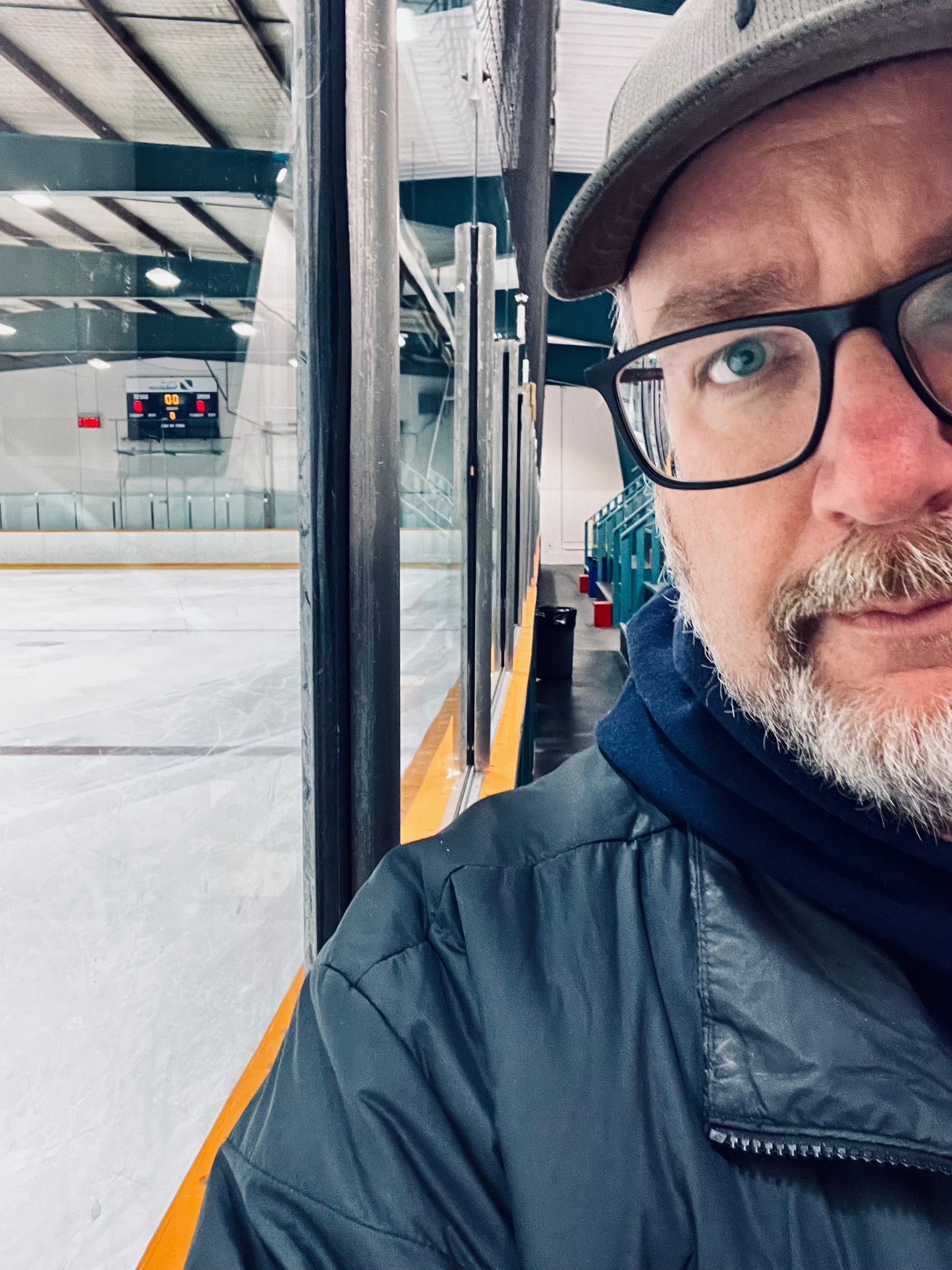 A person wearing glasses and a cap is at an ice skating rink, with a scoreboard visible in the background.
