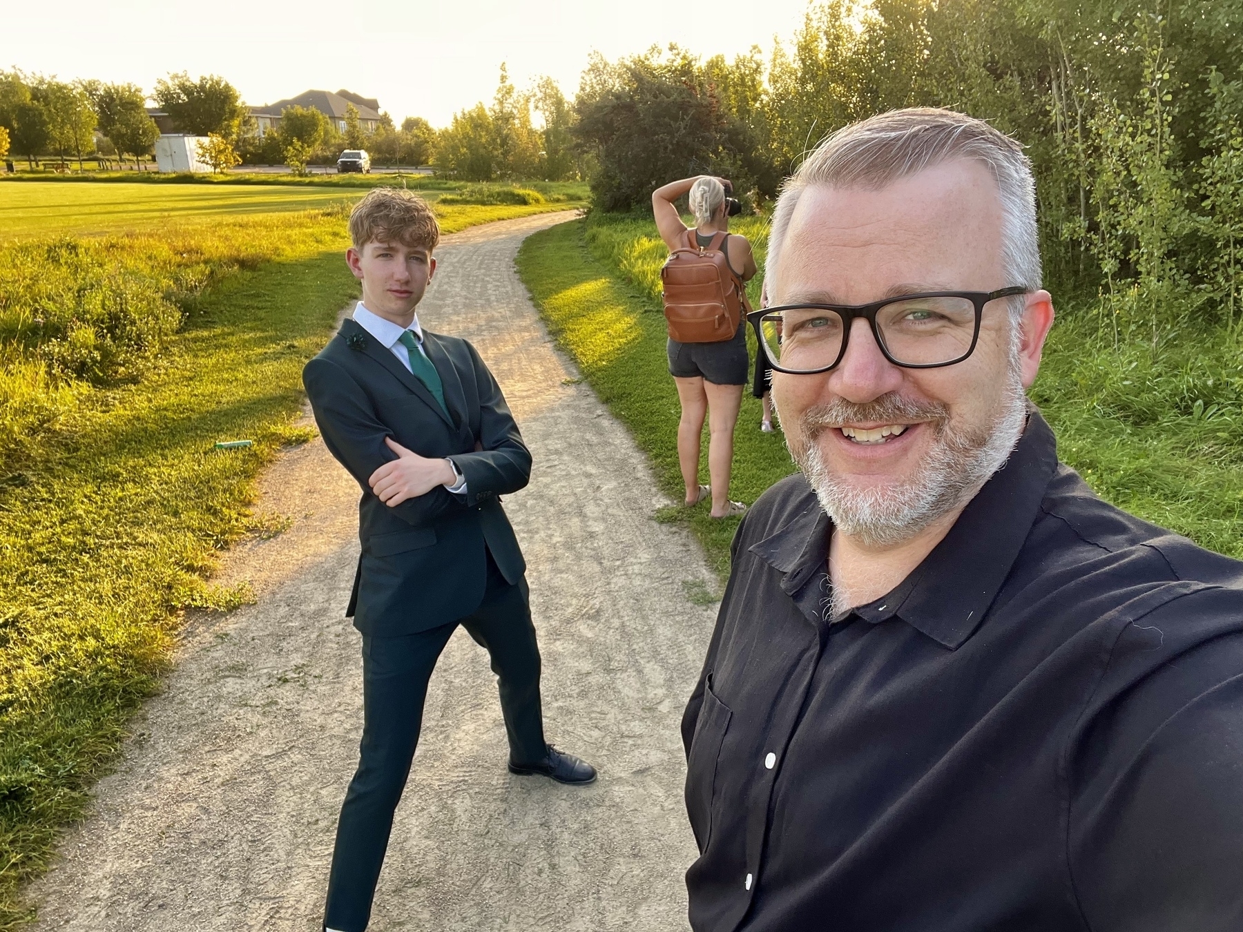 Two people are posing for a selfie on a sunny park path, while a woman in the background is taking pictures. 