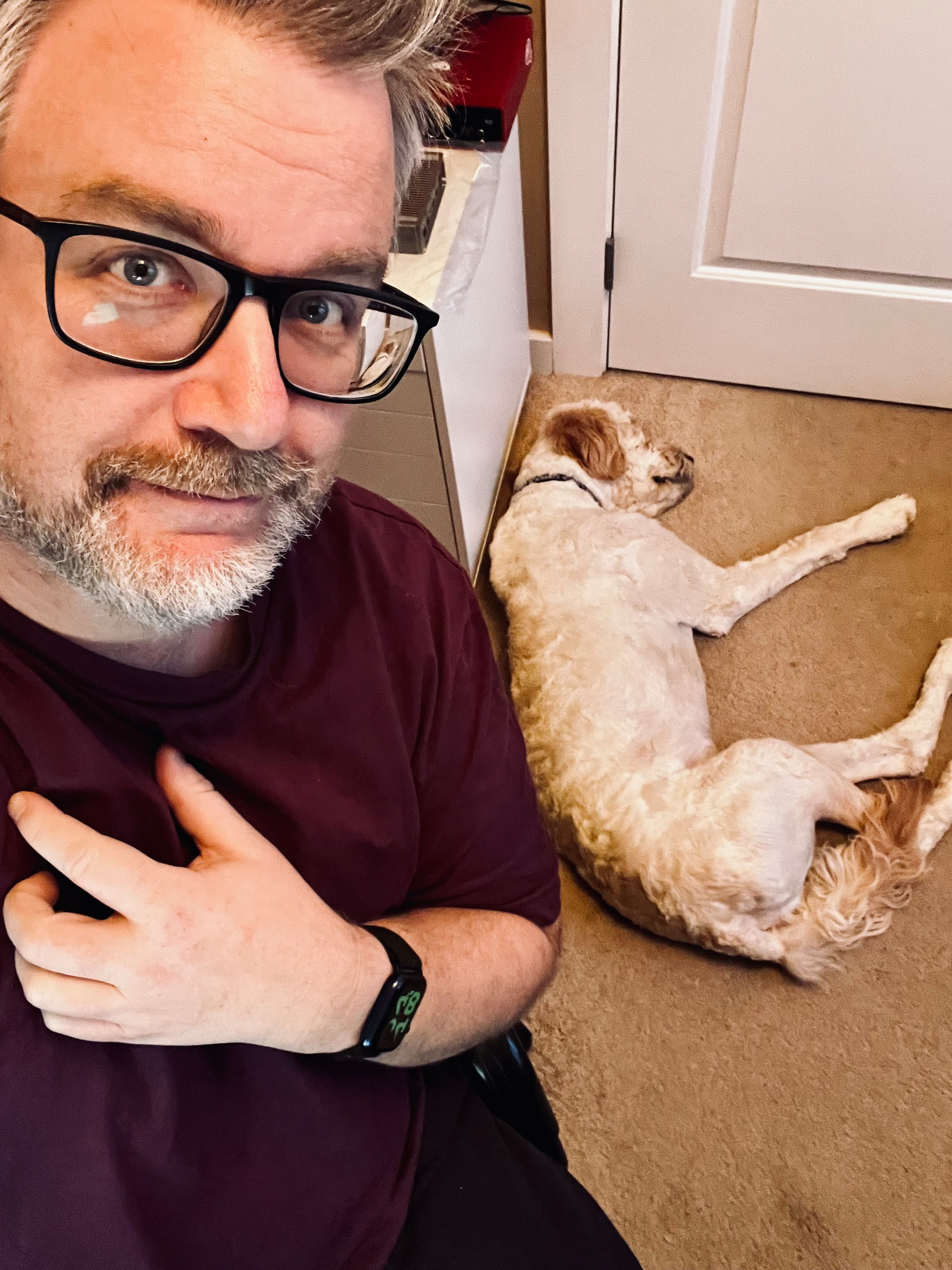 A person with glasses and a smartwatch poses for a selfie while a dog sleeps on the floor behind them.