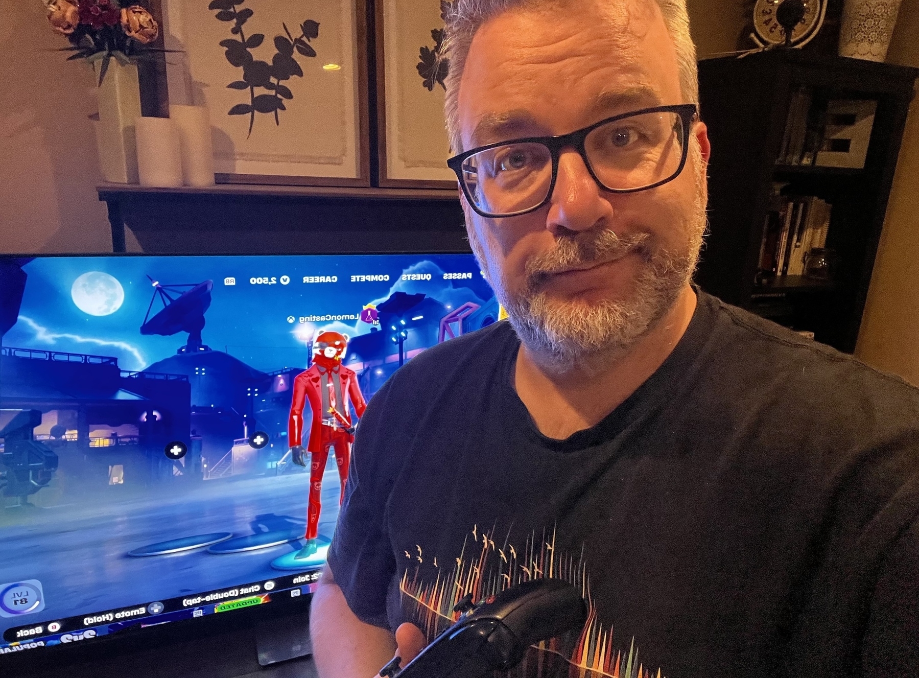 A person wearing glasses and holding a Xbox controller stands in front of a screen displaying a video game character in Fortnite. Lemoncasting is my gamertag in Fortnite.