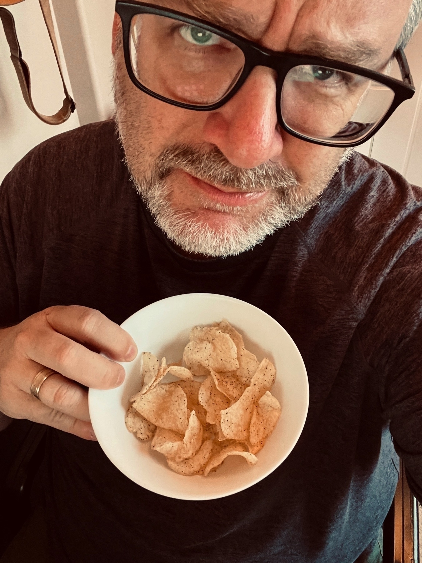 A person with glasses holds a bowl of potato chips while making a facial expression suggesting curiosity or confusion.