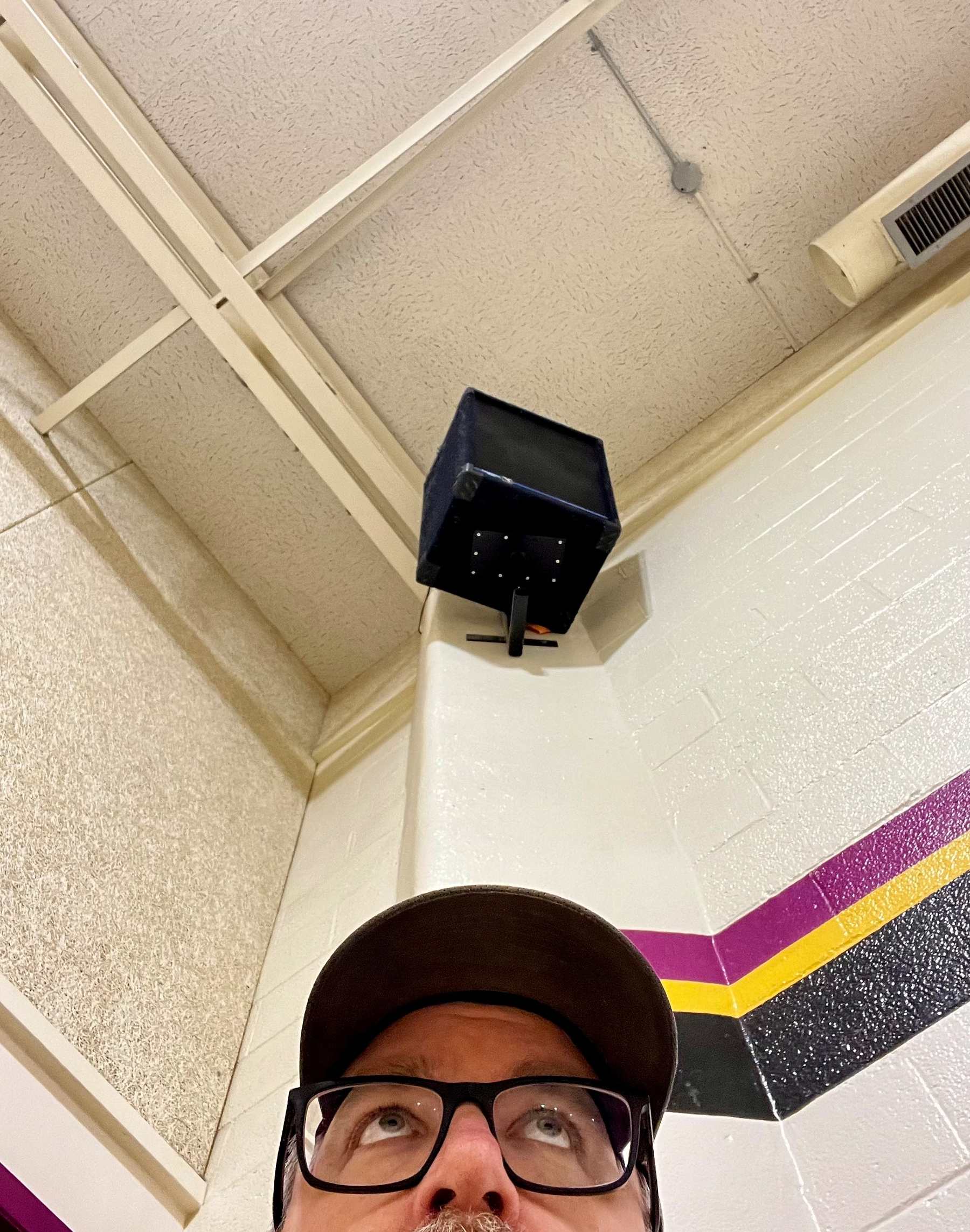 A person wearing a baseball cap is looking up at a mounted black speaker and air vent on a ceiling with a color-striped wall.