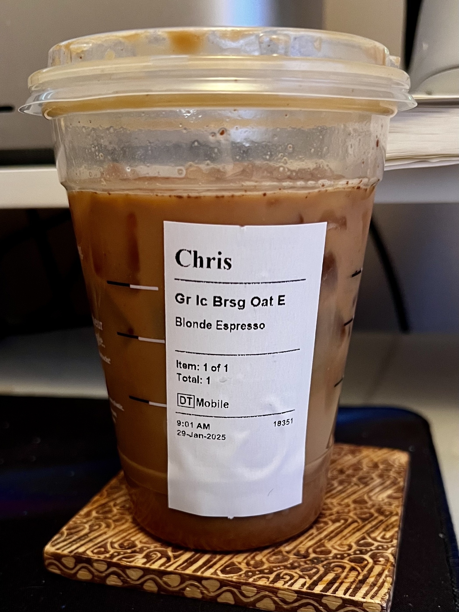 A plastic cup containing an iced coffee beverage with a label showing the order details is placed on a coaster.