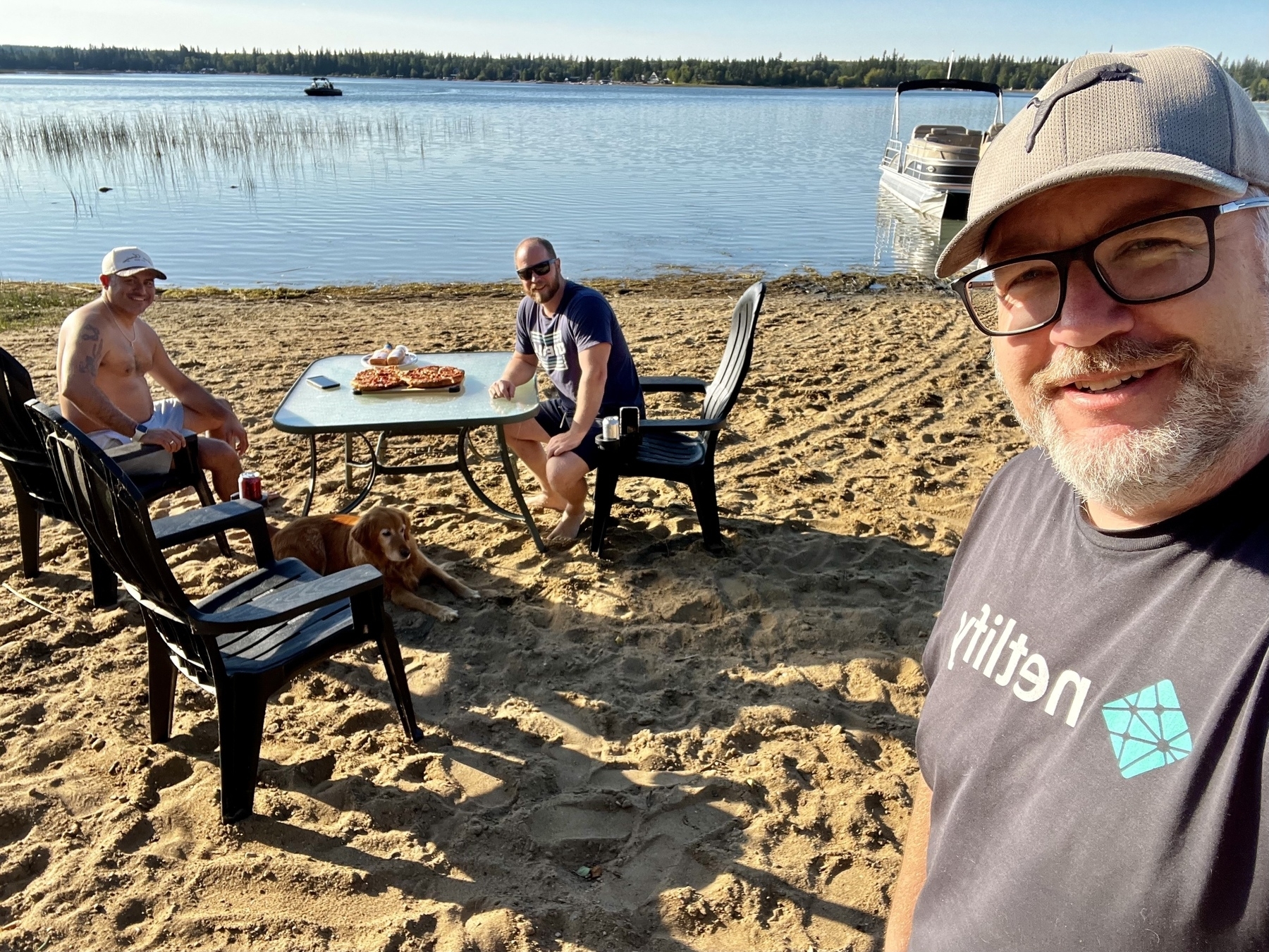 Three men and a dog relax by a lakeside on a sandy beach with a table of food.