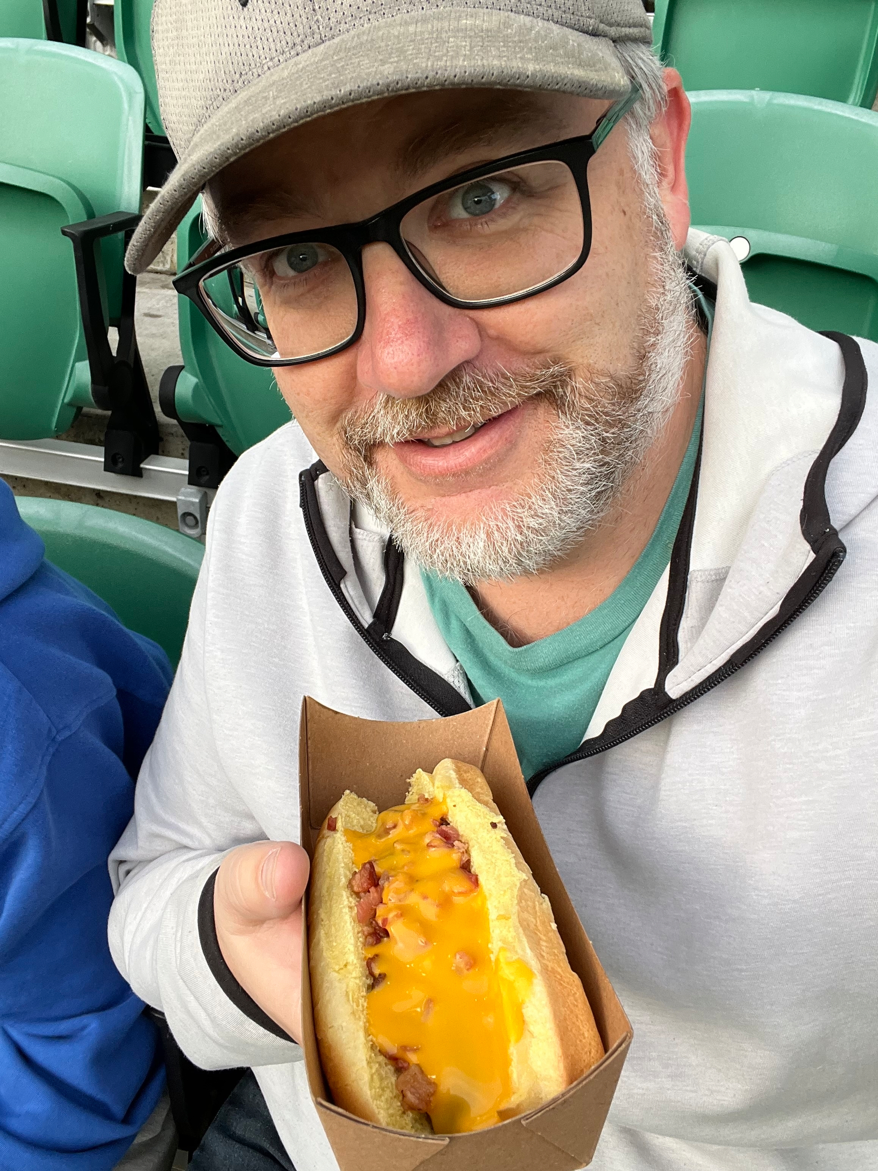 A man wearing glasses and a cap is holding a hot dog topped with cheese and bacon in a cardboard tray.