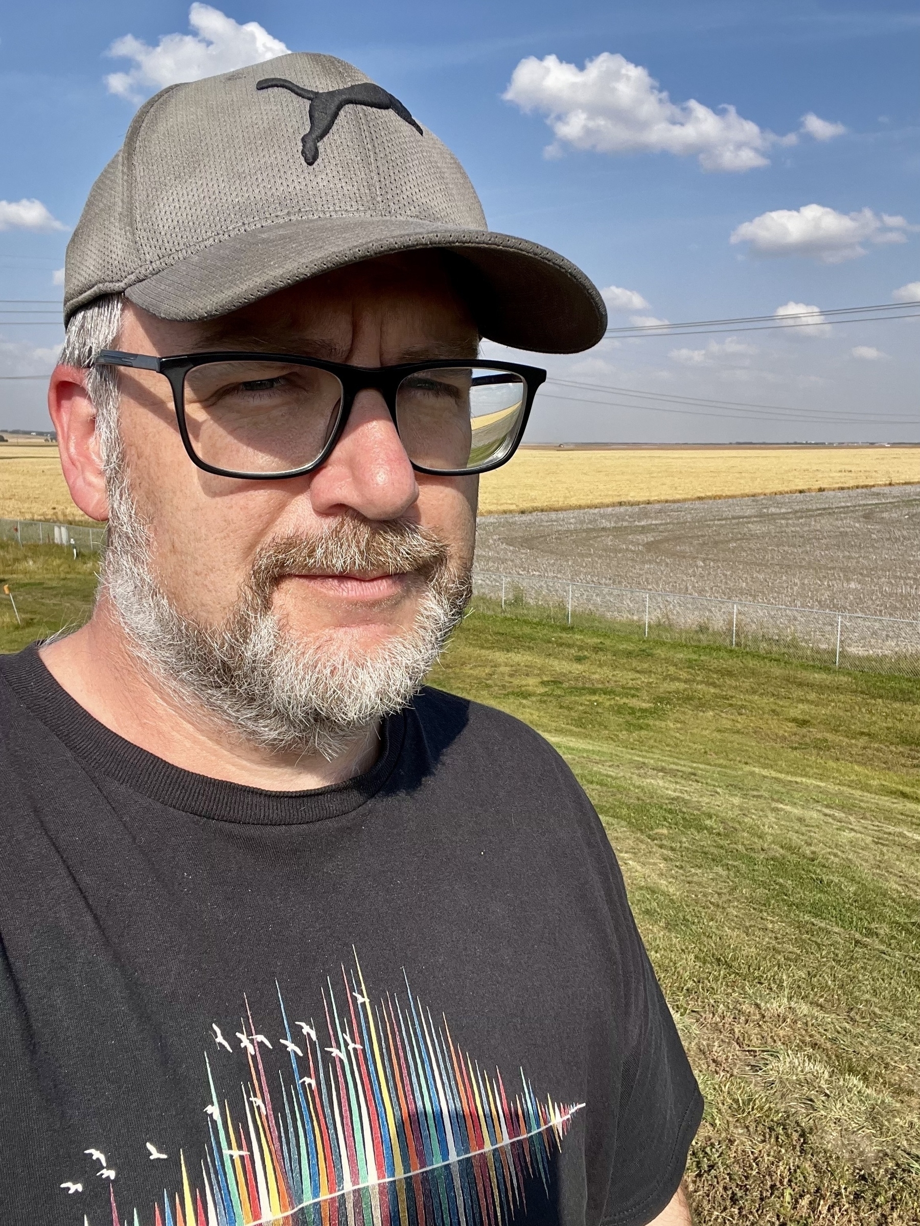 A bearded man wearing a cap and glasses stands in a field under a blue sky with scattered clouds.