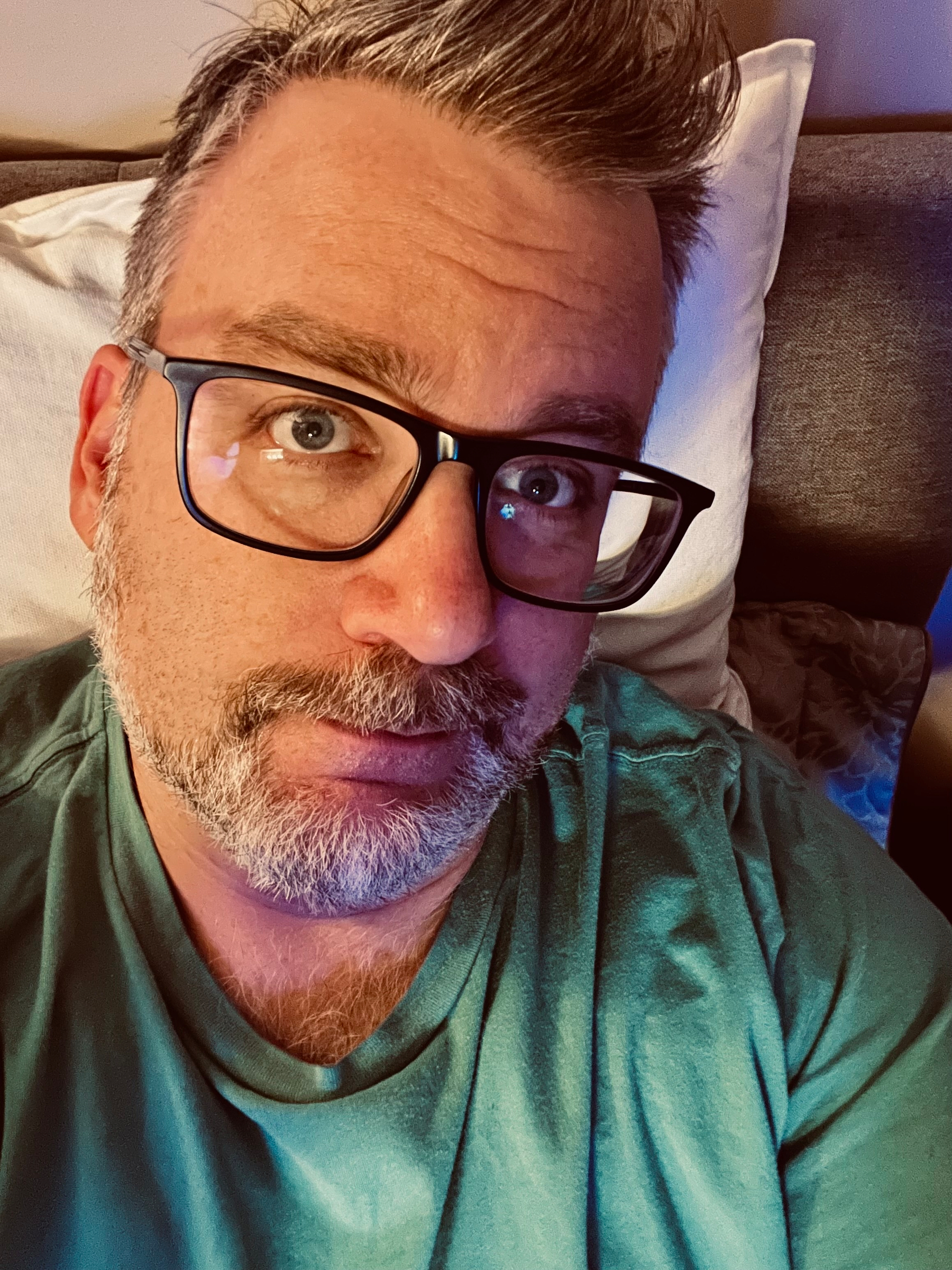 A person with glasses and facial hair is wearing a green shirt while lying on a bed.