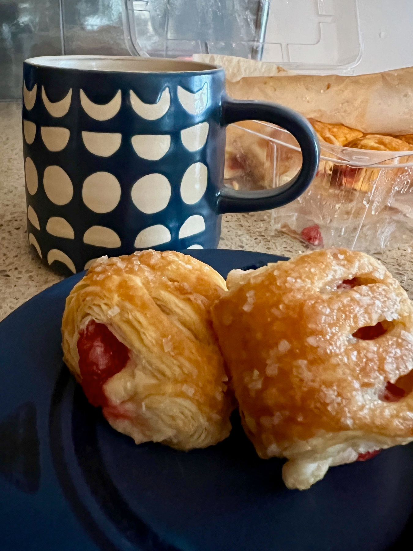 A mug with a geometric pattern is placed beside a blue plate holding two pastries filled with a red fruit filling.