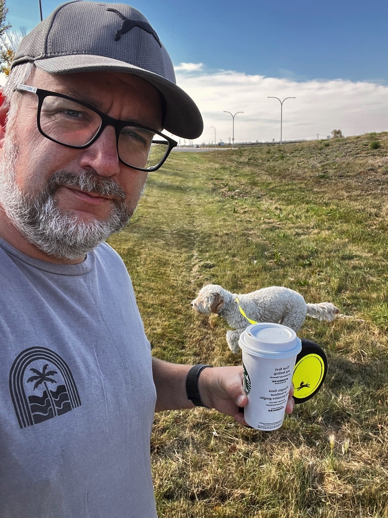A man wearing a cap holds a cup of coffee while a dog walks on a grassy field behind him.