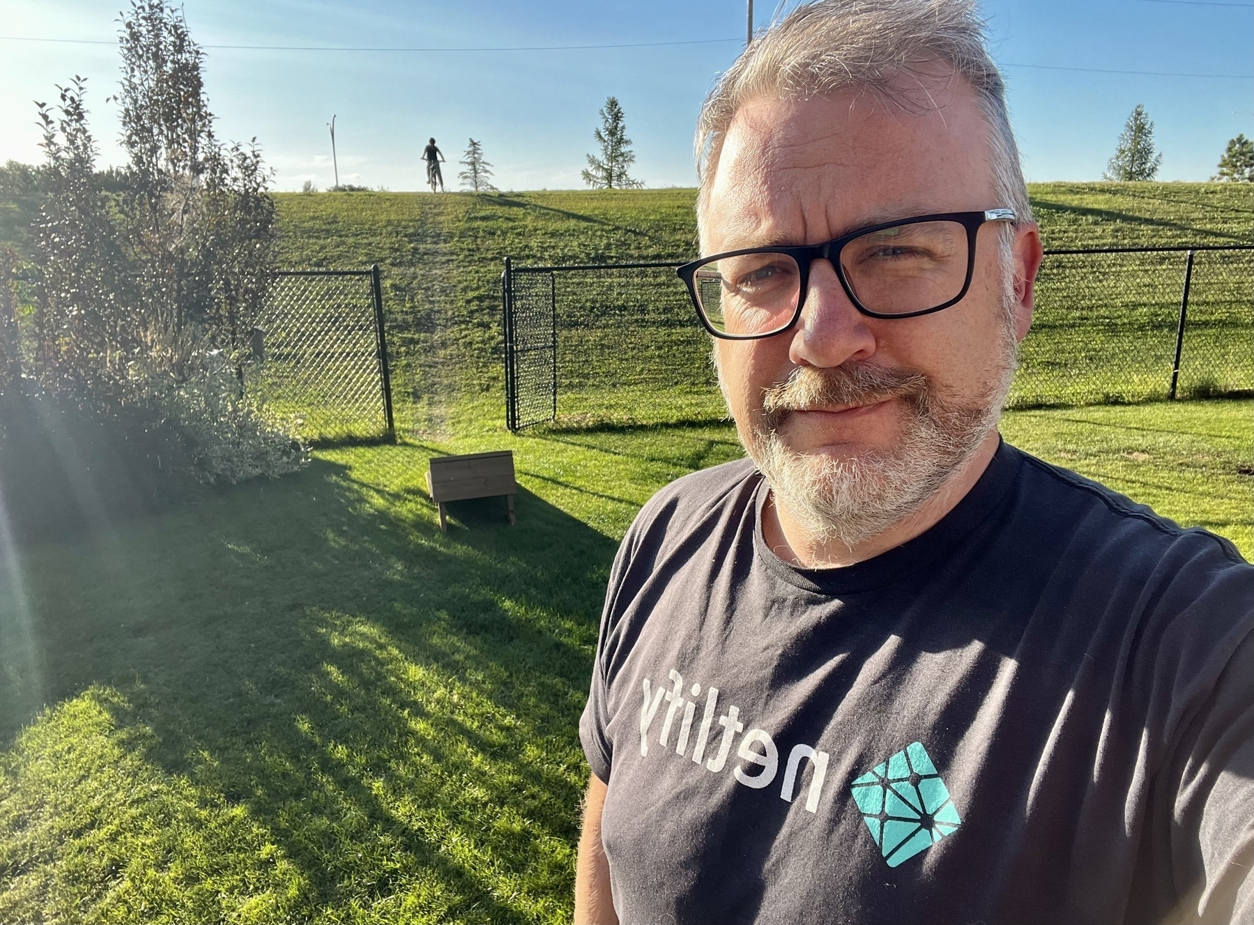 A person wearing glasses and a "Netlify" t-shirt stands outside on a grassy lawn with a wire fence and some trees in the background.