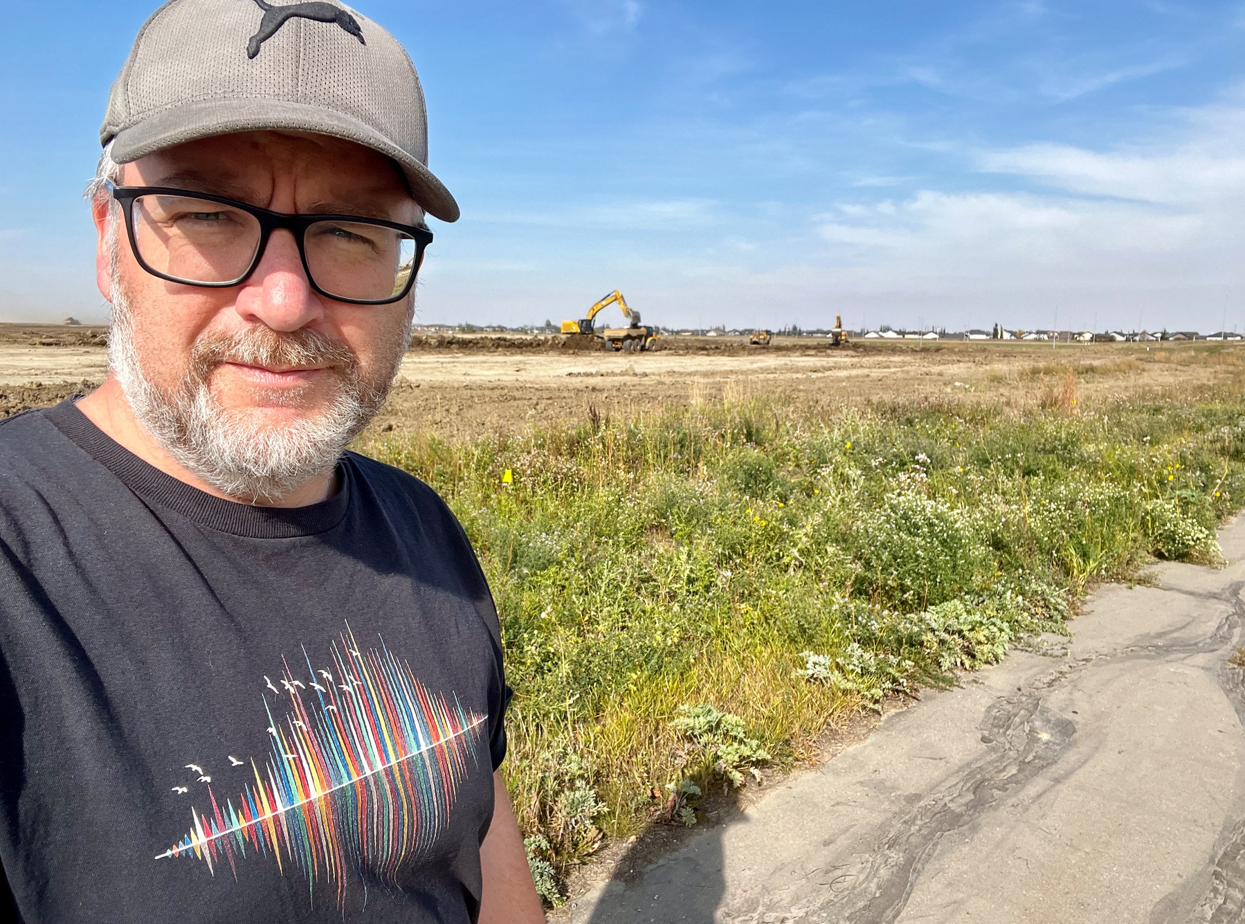 Seflie of me with a hat, beard, and glasses standing in front of a field with a dump truck and diggers clearing and moving dirt around.