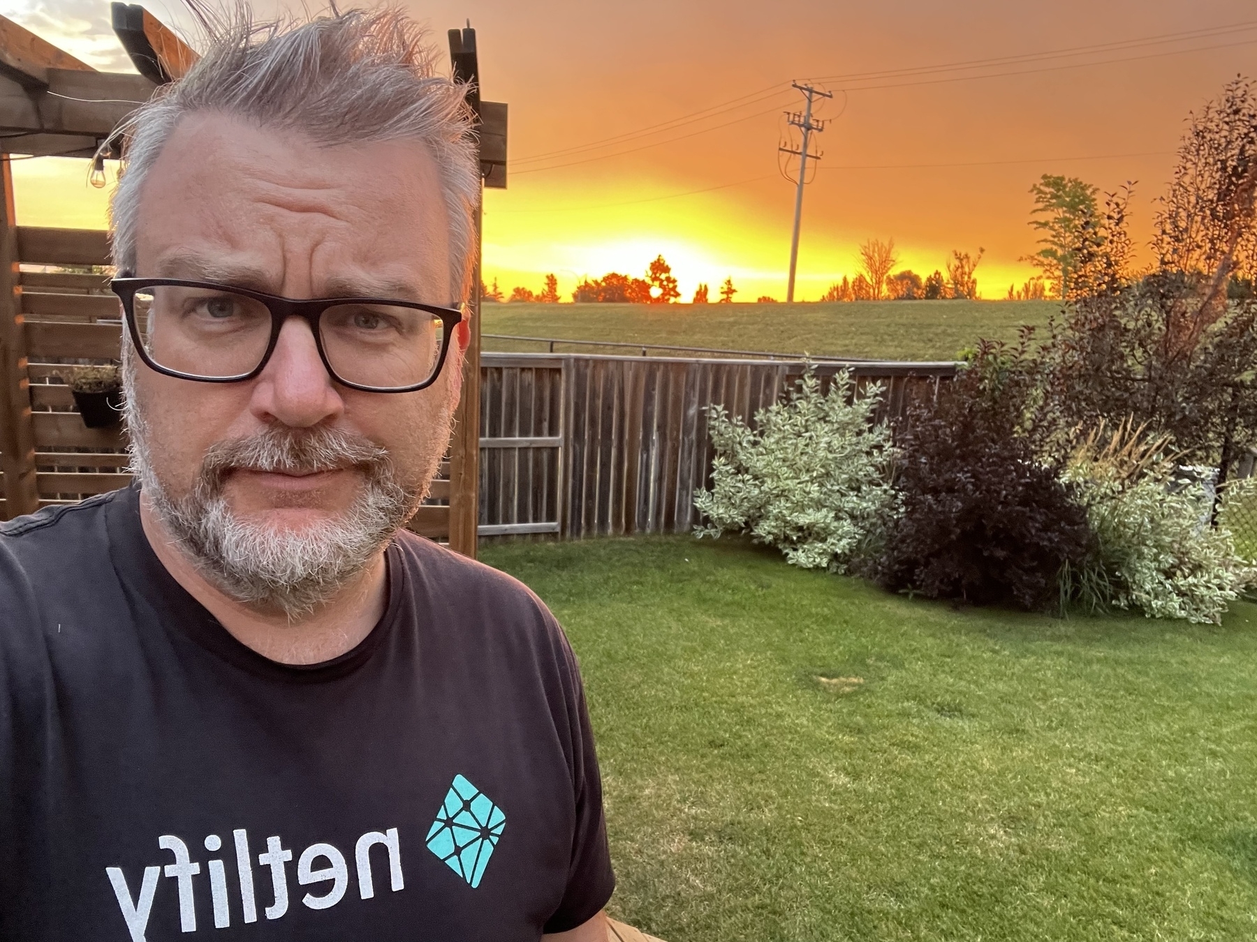 A person with glasses stands in a backyard at sunset, with a wooden fence and lush greenery in the background.