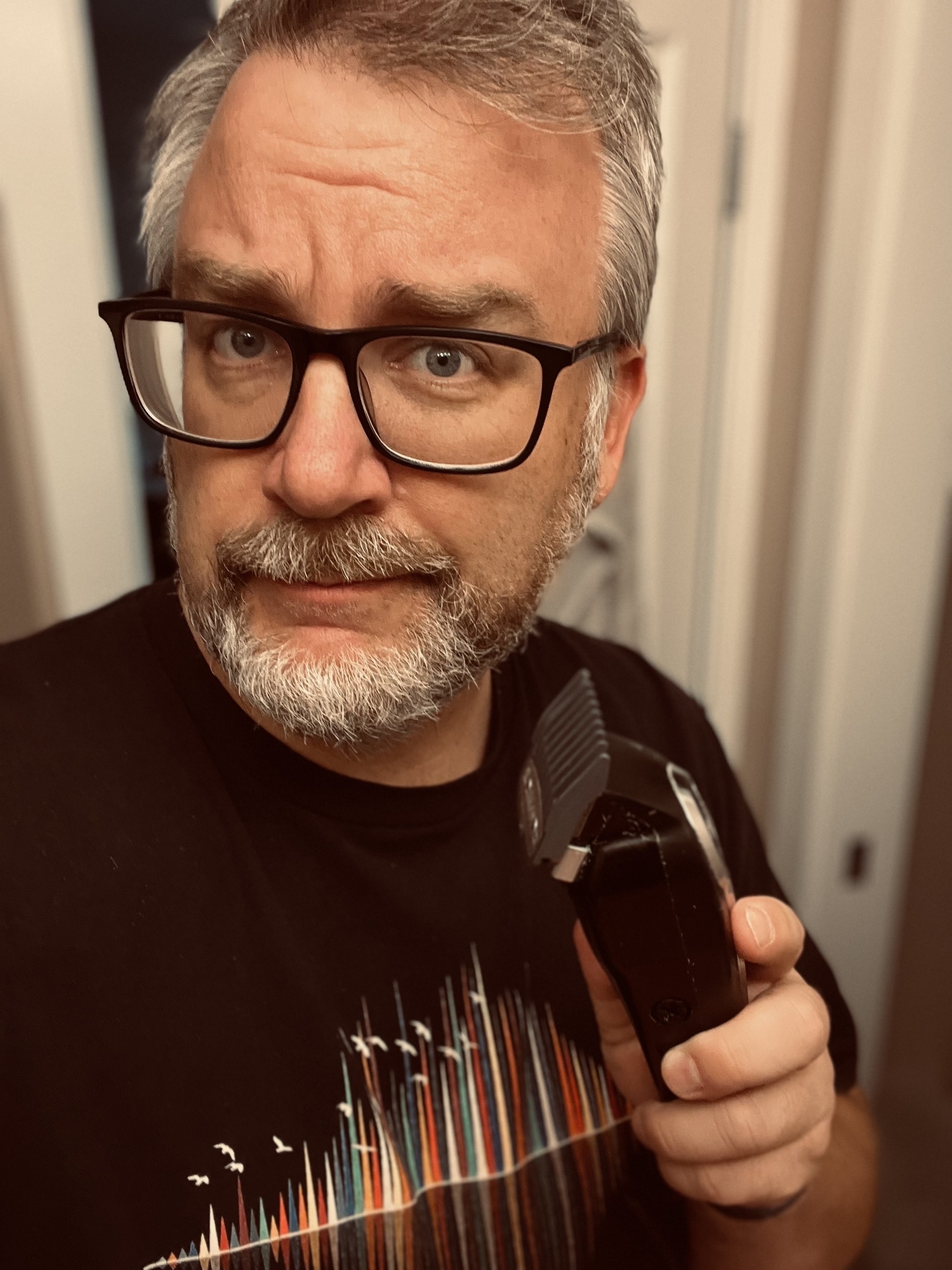 A person with glasses and a beard holds an electric hair clipper, standing indoors.