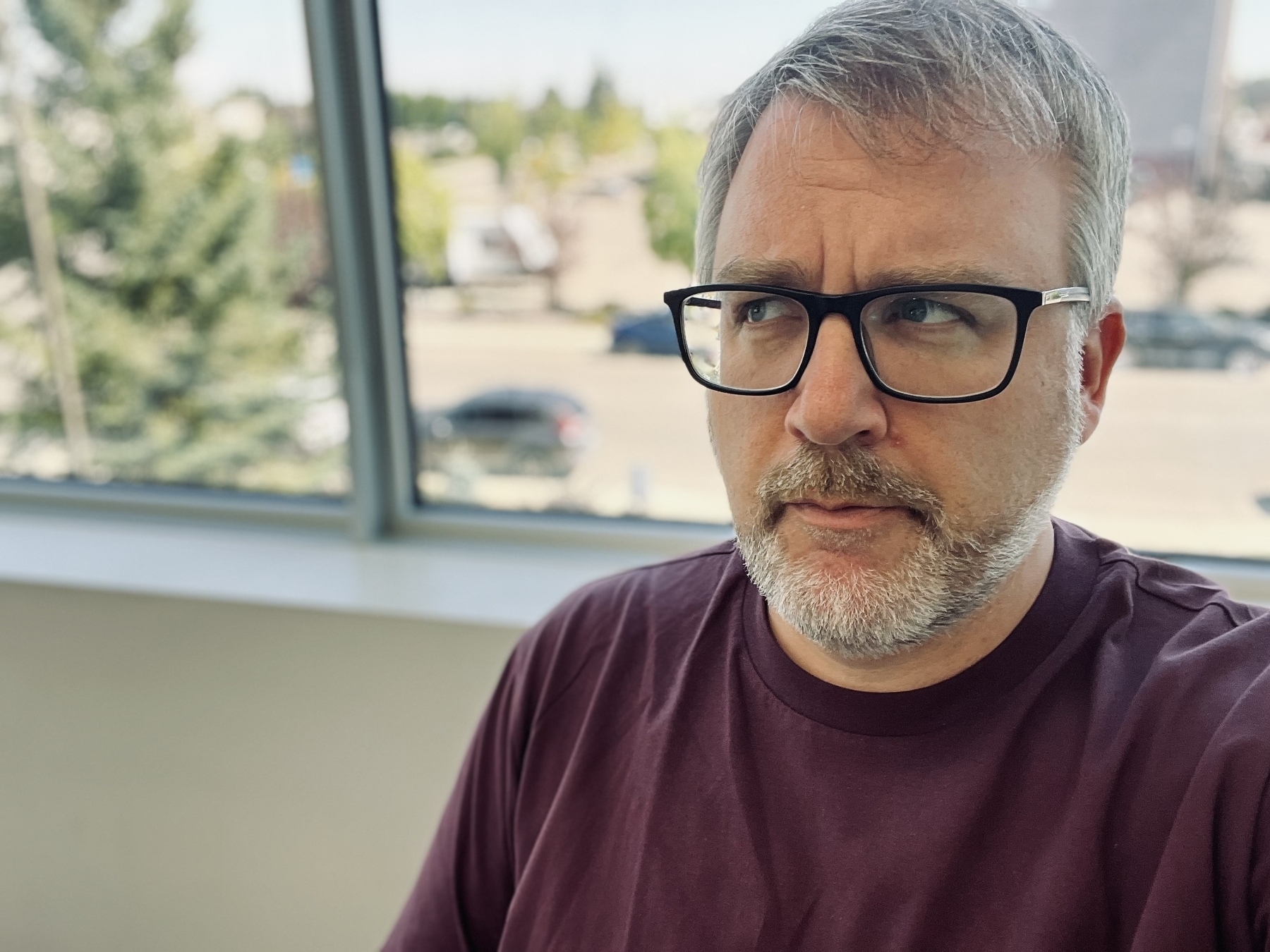 A man with glasses and a beard is looking thoughtfully to the side while standing indoors near a window.