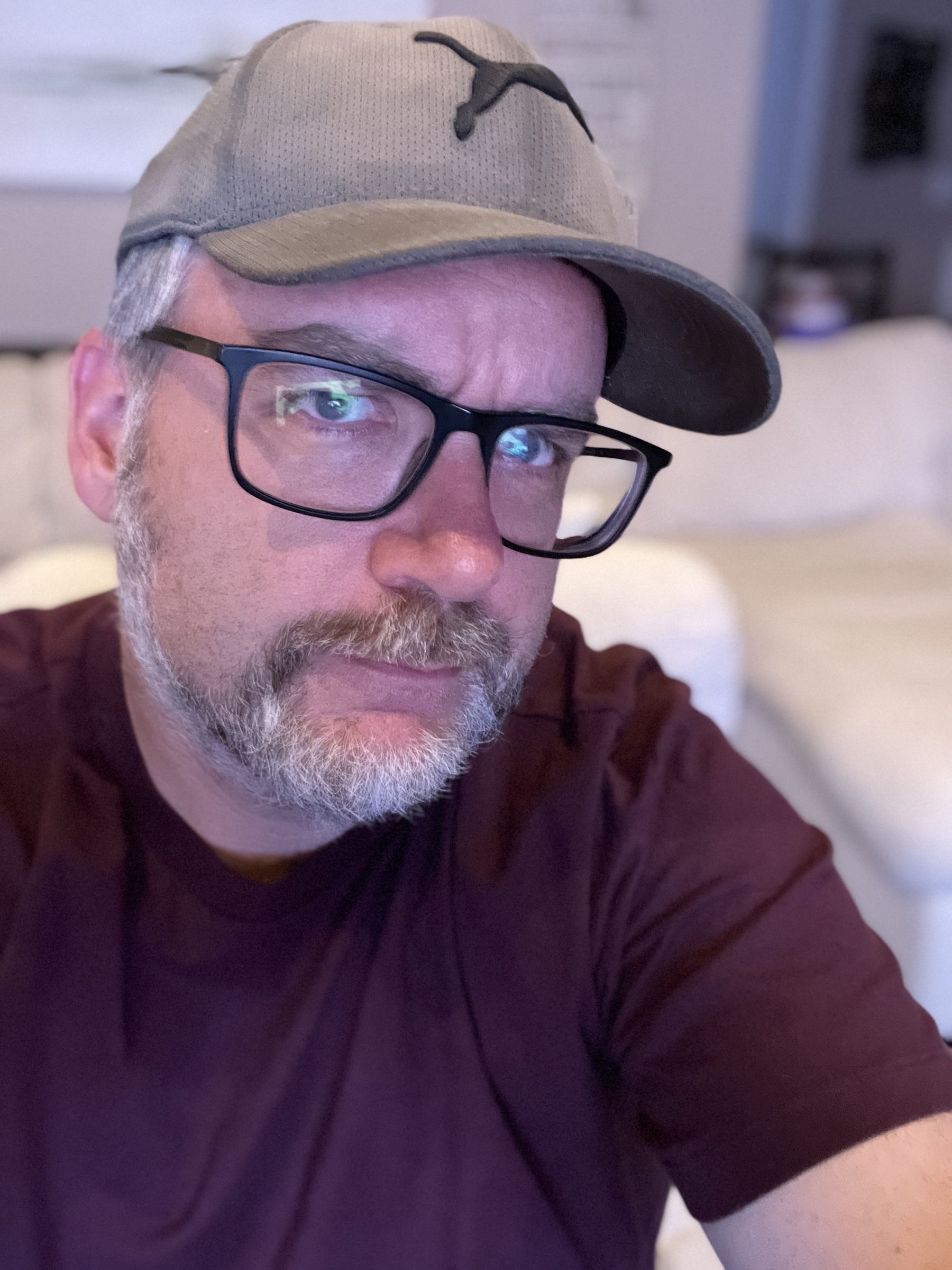 A man wearing glasses and a cap is taking a selfie in a room with a white sofa in the background.